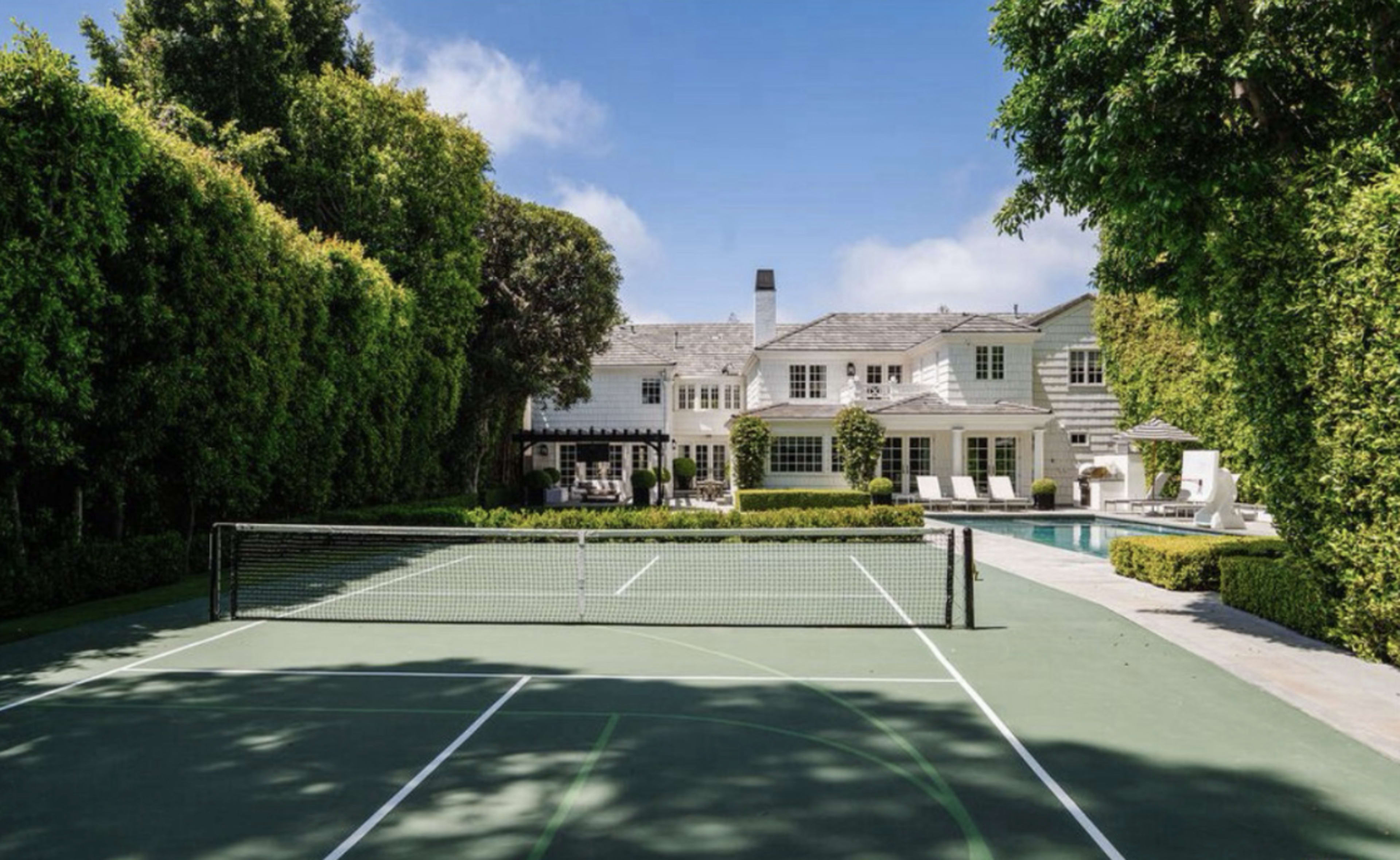 A well-maintained tennis court is positioned in front of a large, multi-level home surrounded by lush greenery and a pool area.