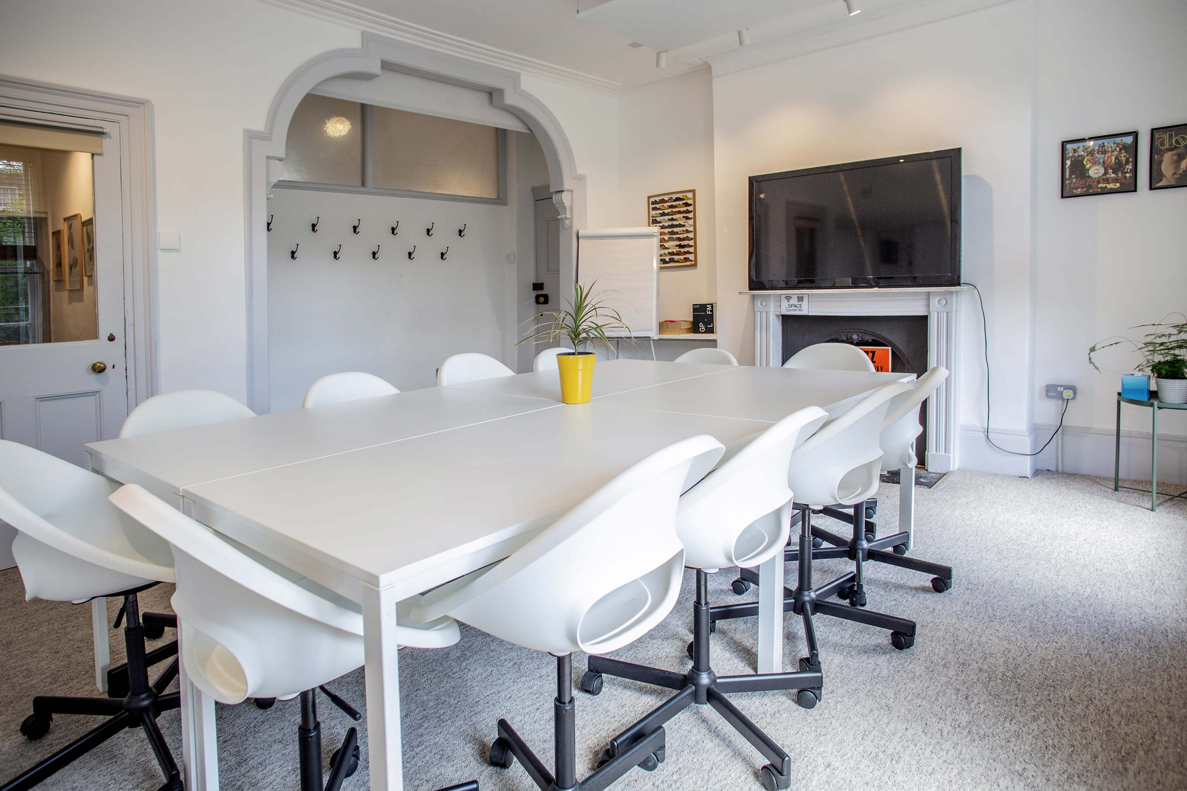 A modern conference room features a large white table surrounded by twelve white chairs, with a television mounted on the wall and a small plant on the side.