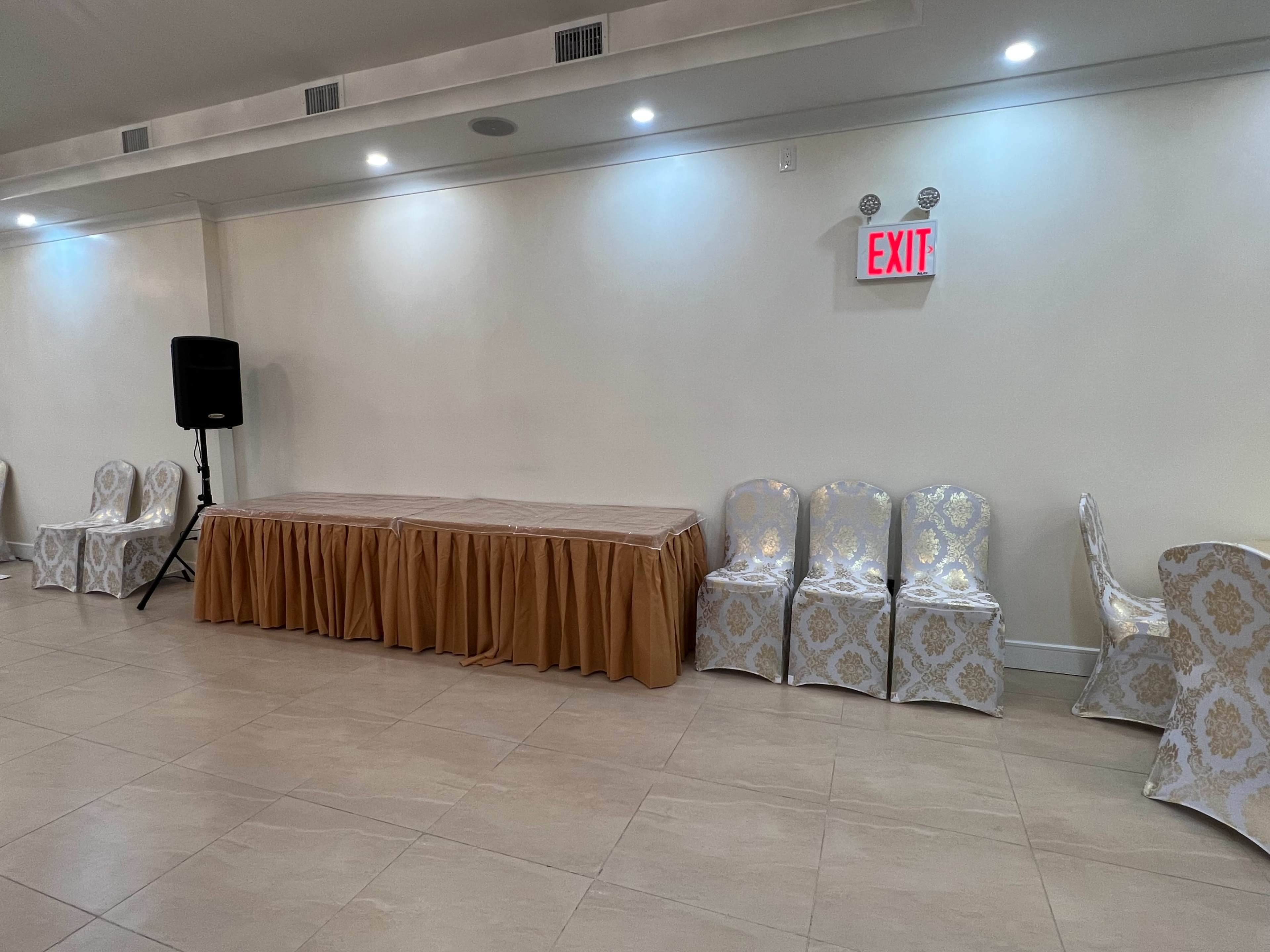 A spacious room with a table covered in a brown tablecloth and several patterned chairs arranged alongside a wall, featuring an exit sign and a speaker in the corner.