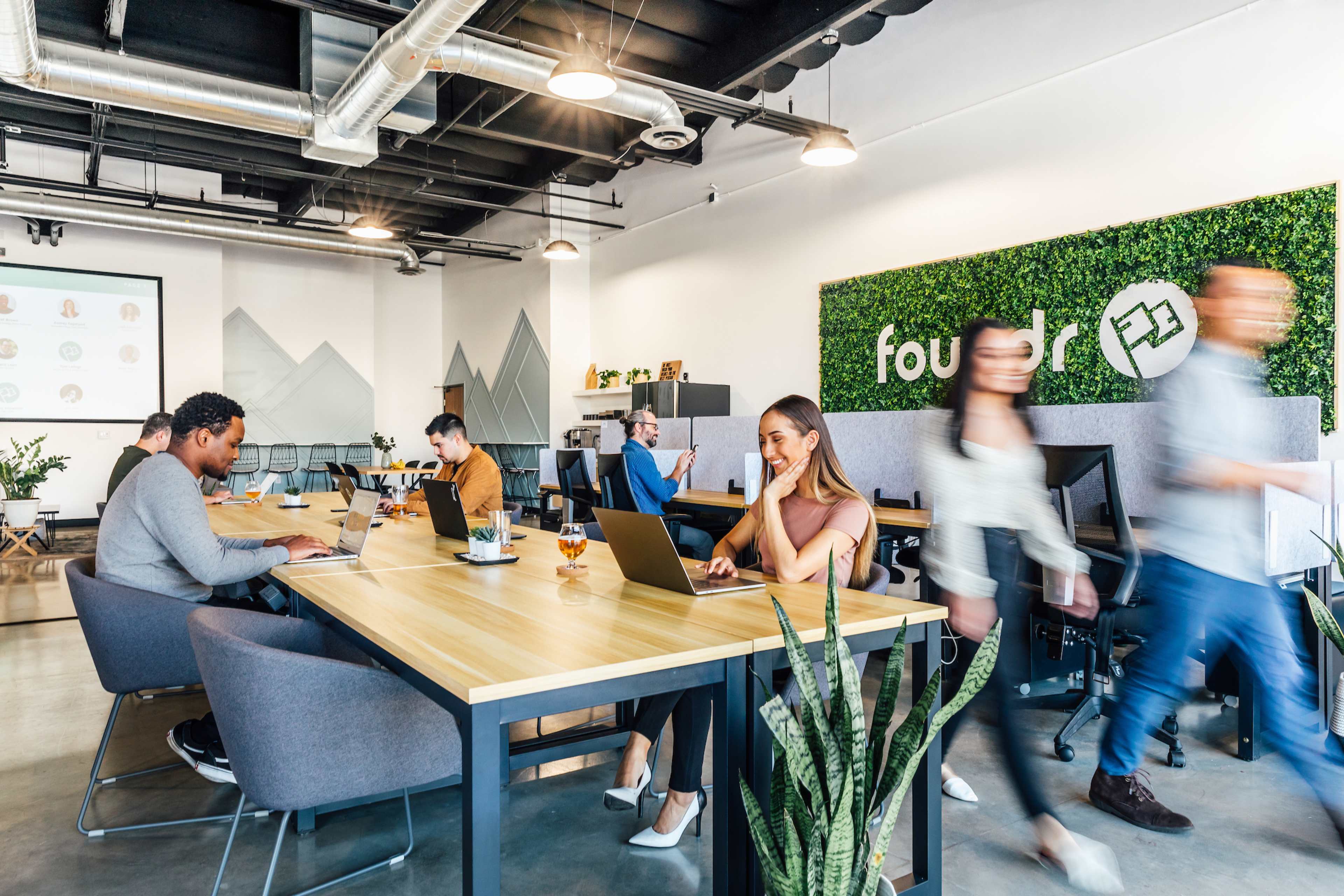 A modern open office space with several people working on laptops at wooden tables, a large screen displaying information, and greenery on the walls.