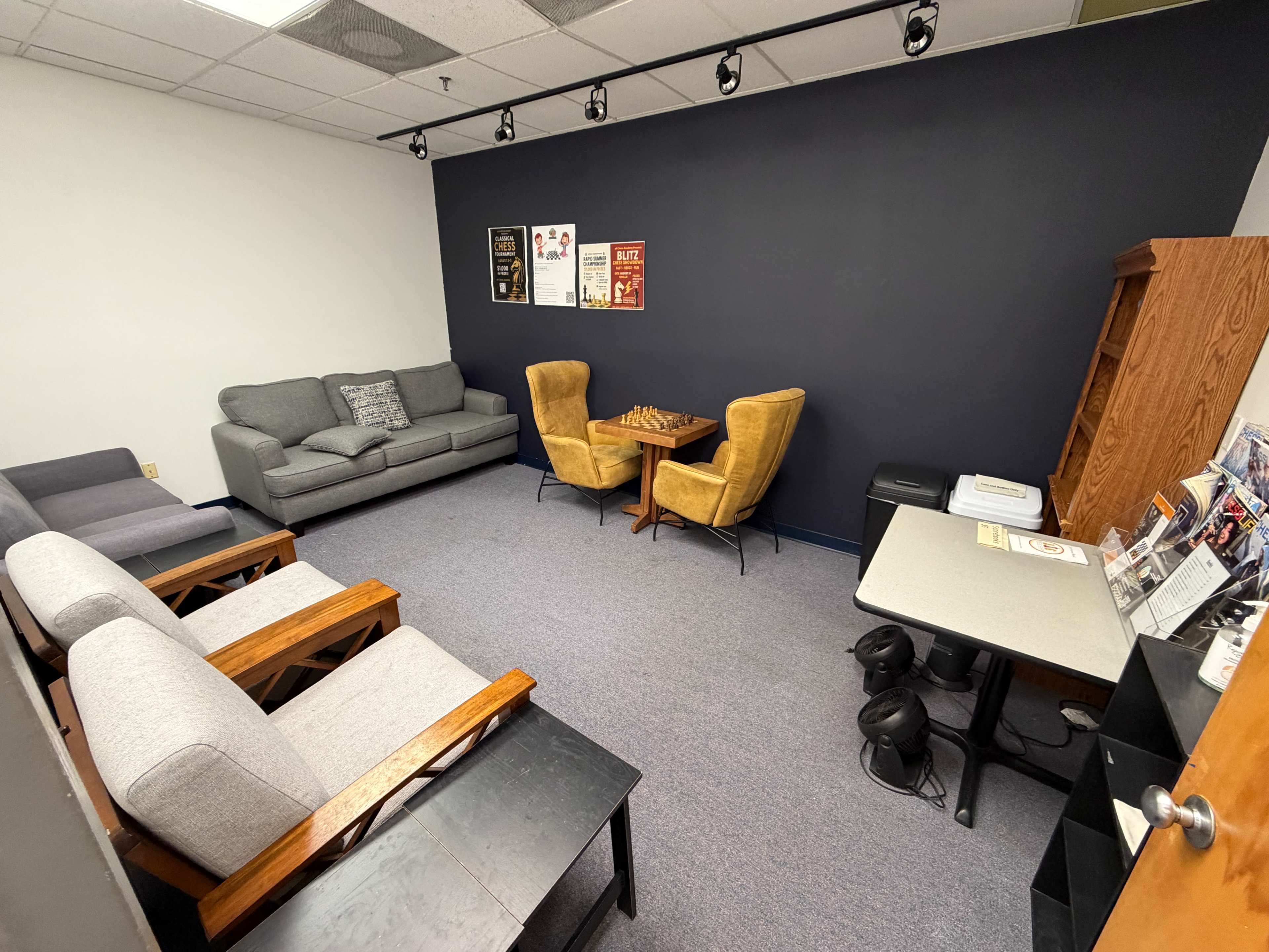 The image shows a small lounge area with gray sofas, two yellow armchairs, a chessboard, and a table with magazines against a dark wall.