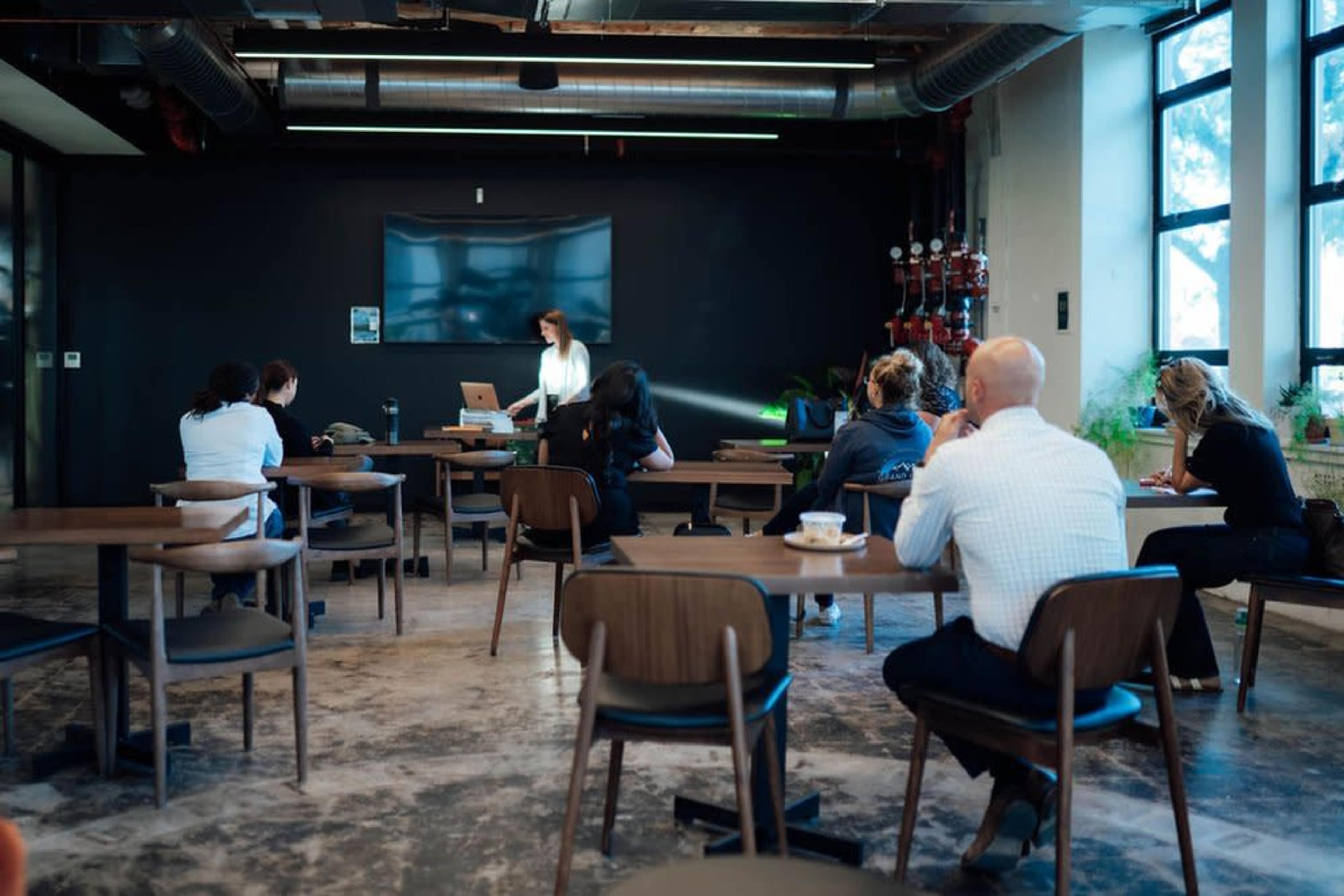 A woman presents in front of a small audience seated at tables in a modern interior space with large windows.