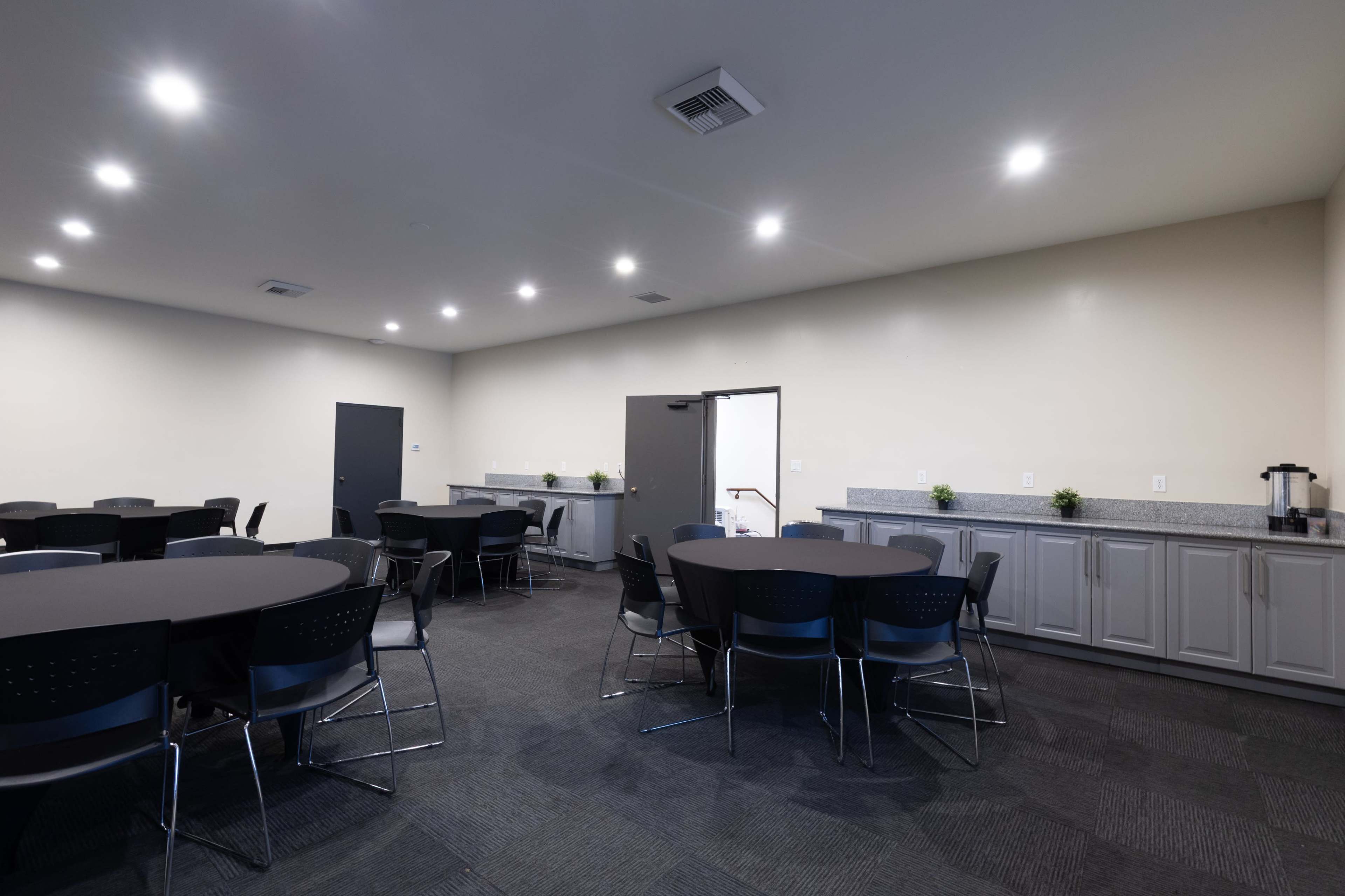 The image shows a well-lit meeting room containing several round tables and chairs arranged neatly, with a countertop area and a door leading to another space.