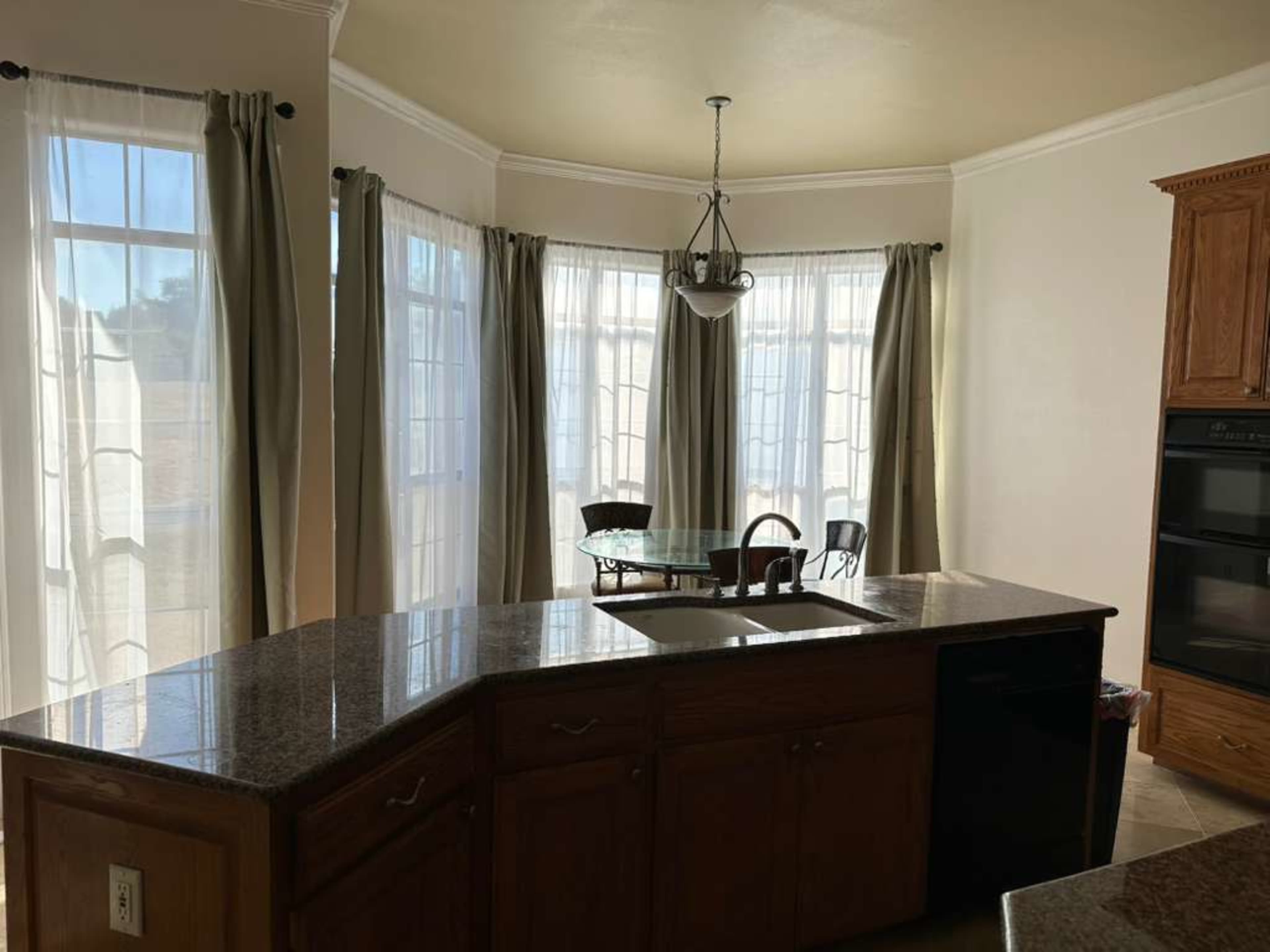 The image shows a kitchen area featuring an island with a sink, a dining table, and large windows draped with sheer curtains.