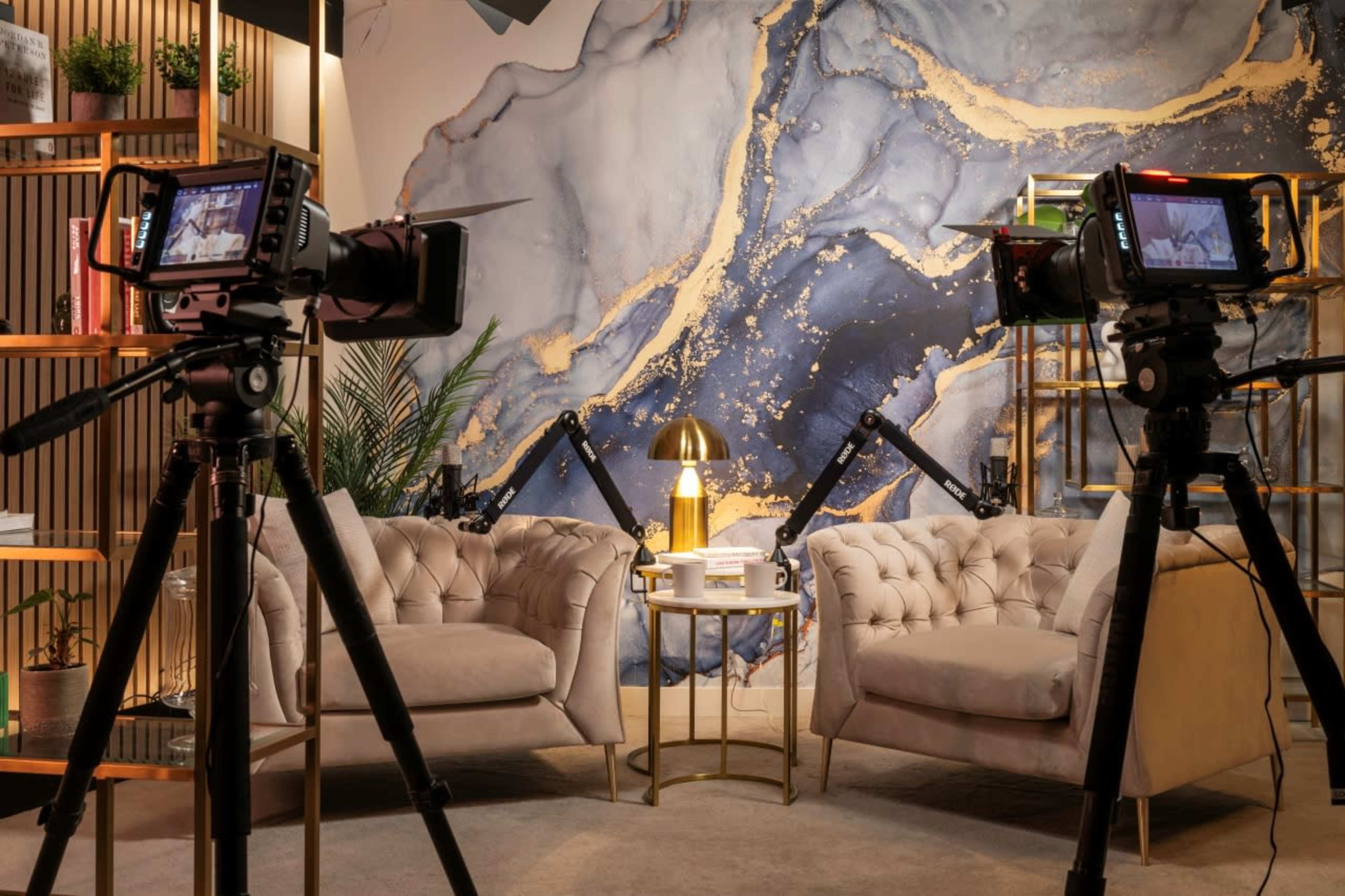 A modern interview setup with two plush sofas facing each other, a small table with decor, and two cameras on tripods capturing the scene against a blue and gold abstract wall.