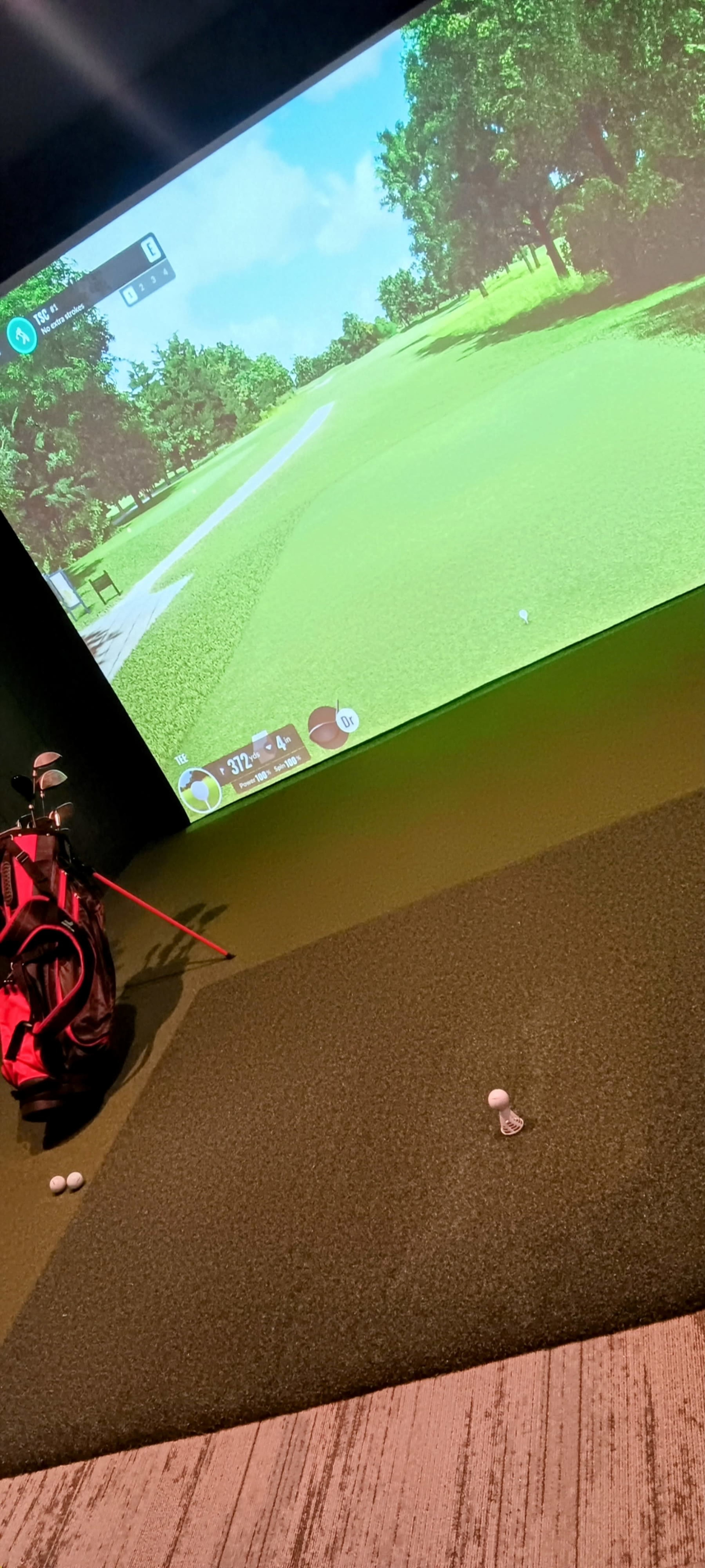 The image shows a golf simulator setup with a digital screen displaying a golf course and a golf bag with clubs beside a mat on the floor where golf balls are positioned.