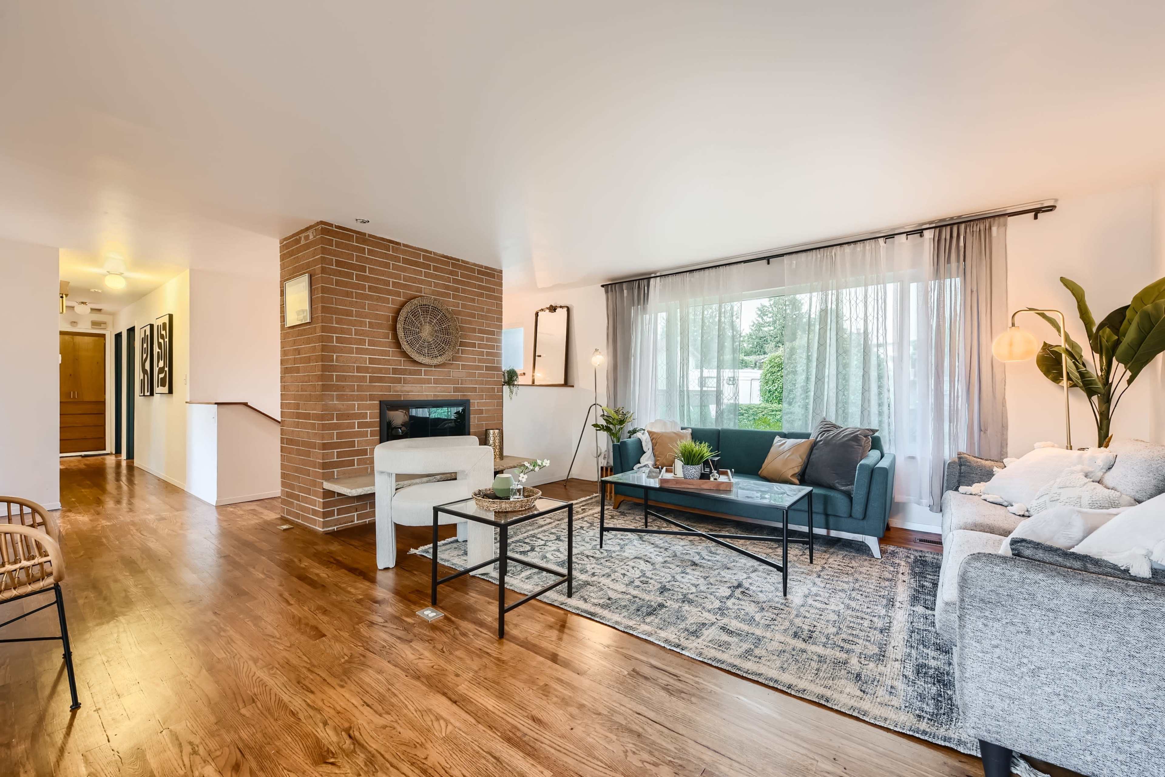 The image shows a modern living room with a brick accent wall, a fireplace, large windows, and a seating arrangement featuring a sofa and armchairs.