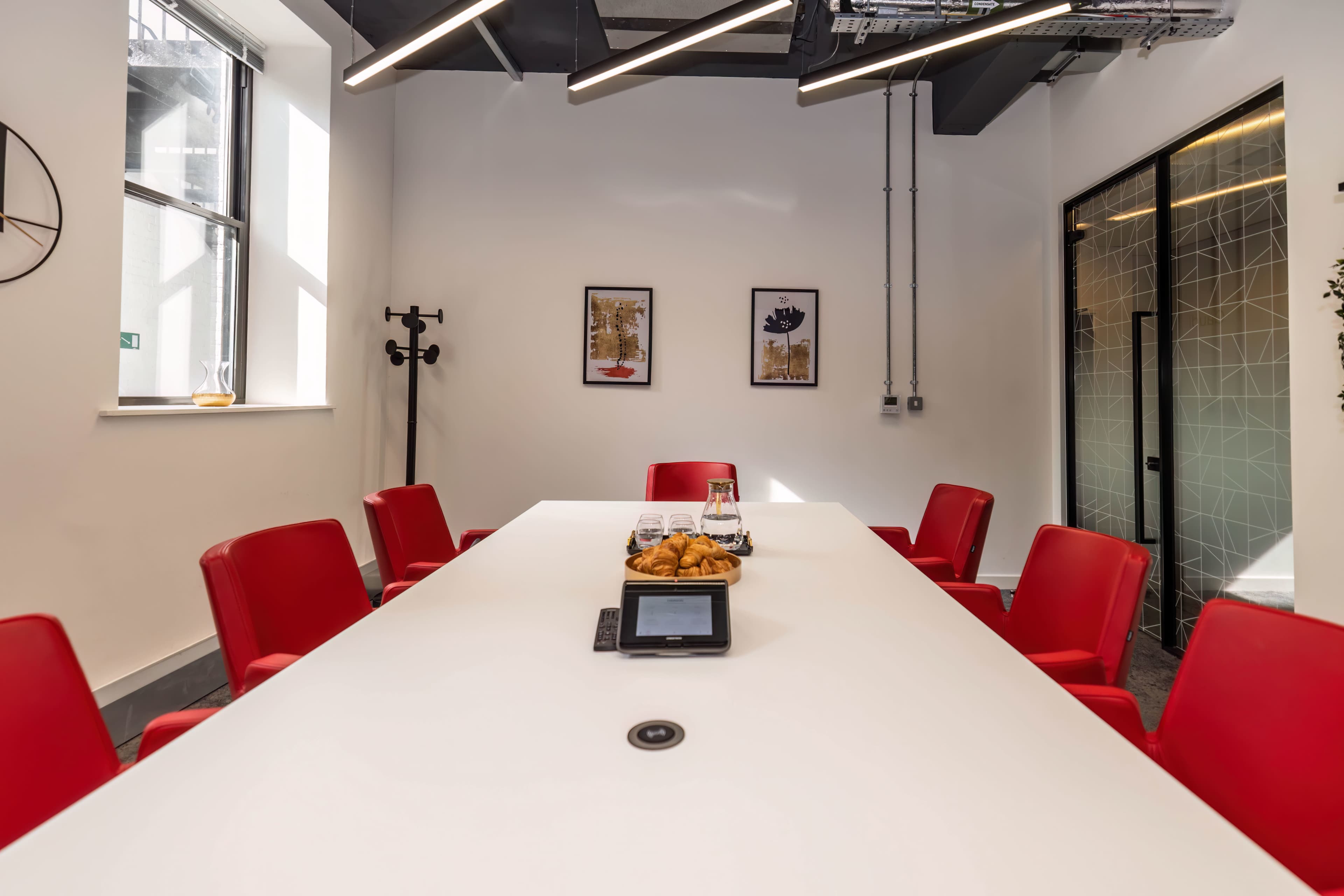 A modern conference room features a white table surrounded by red chairs, with framed artworks on the wall and a tray of pastries on the table.