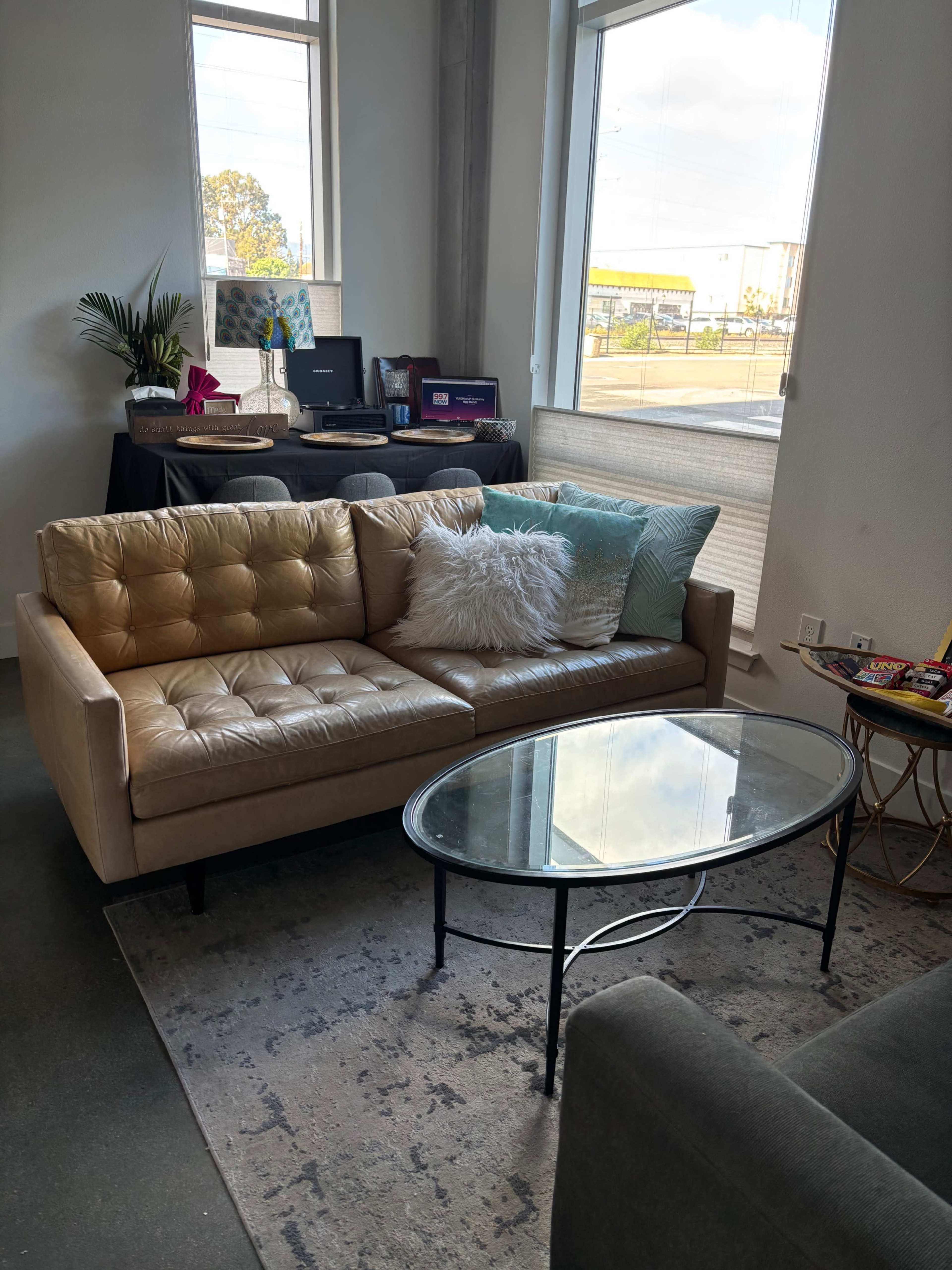 A beige sofa is positioned in a brightly lit room with a glass coffee table in front and a desk in the background.