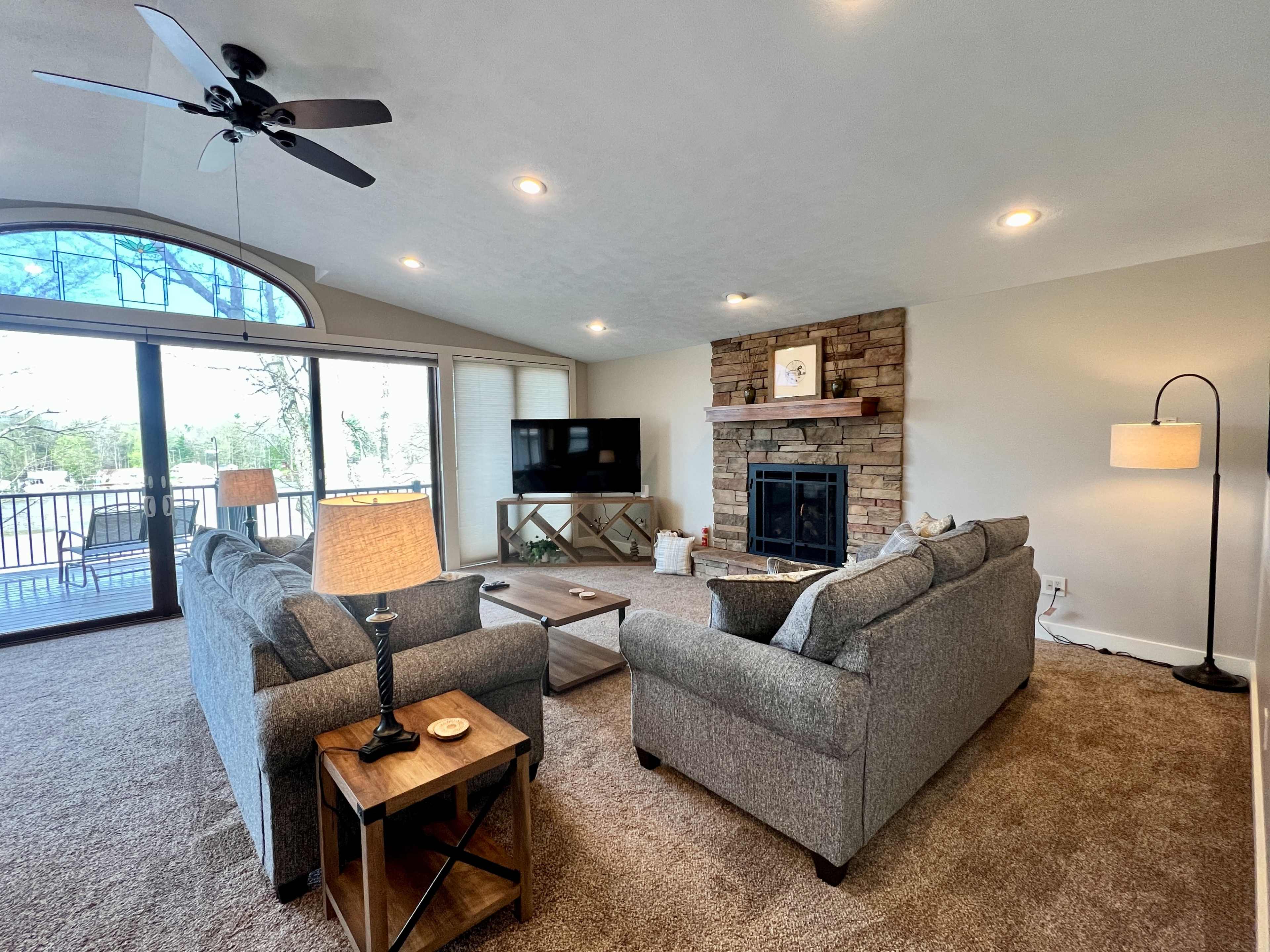 The living room features two gray sofas facing a stone fireplace, with a ceiling fan overhead and large windows allowing natural light.
