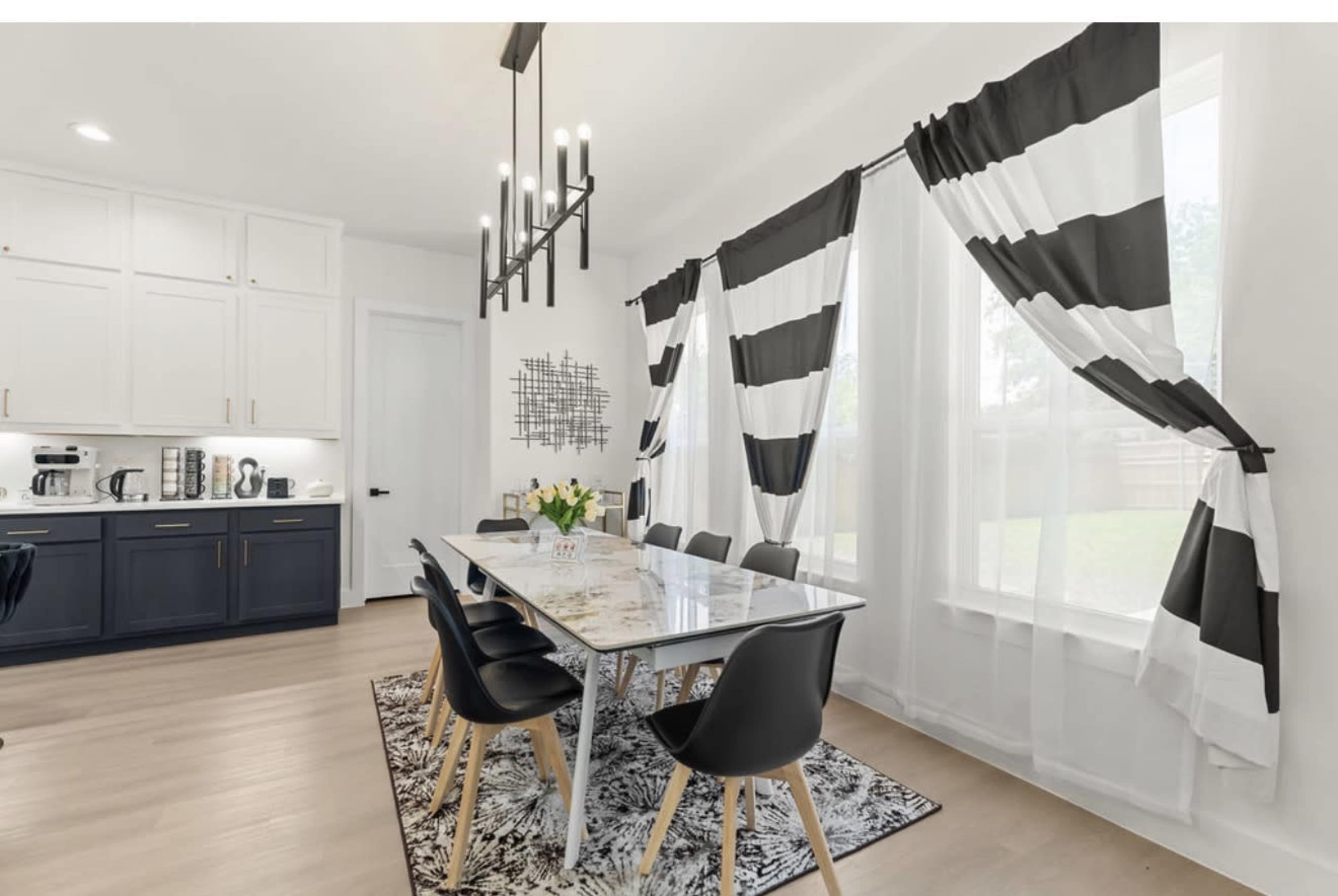 The image shows a modern dining area featuring a glass table surrounded by black chairs, with large windows draped in black and white striped curtains.