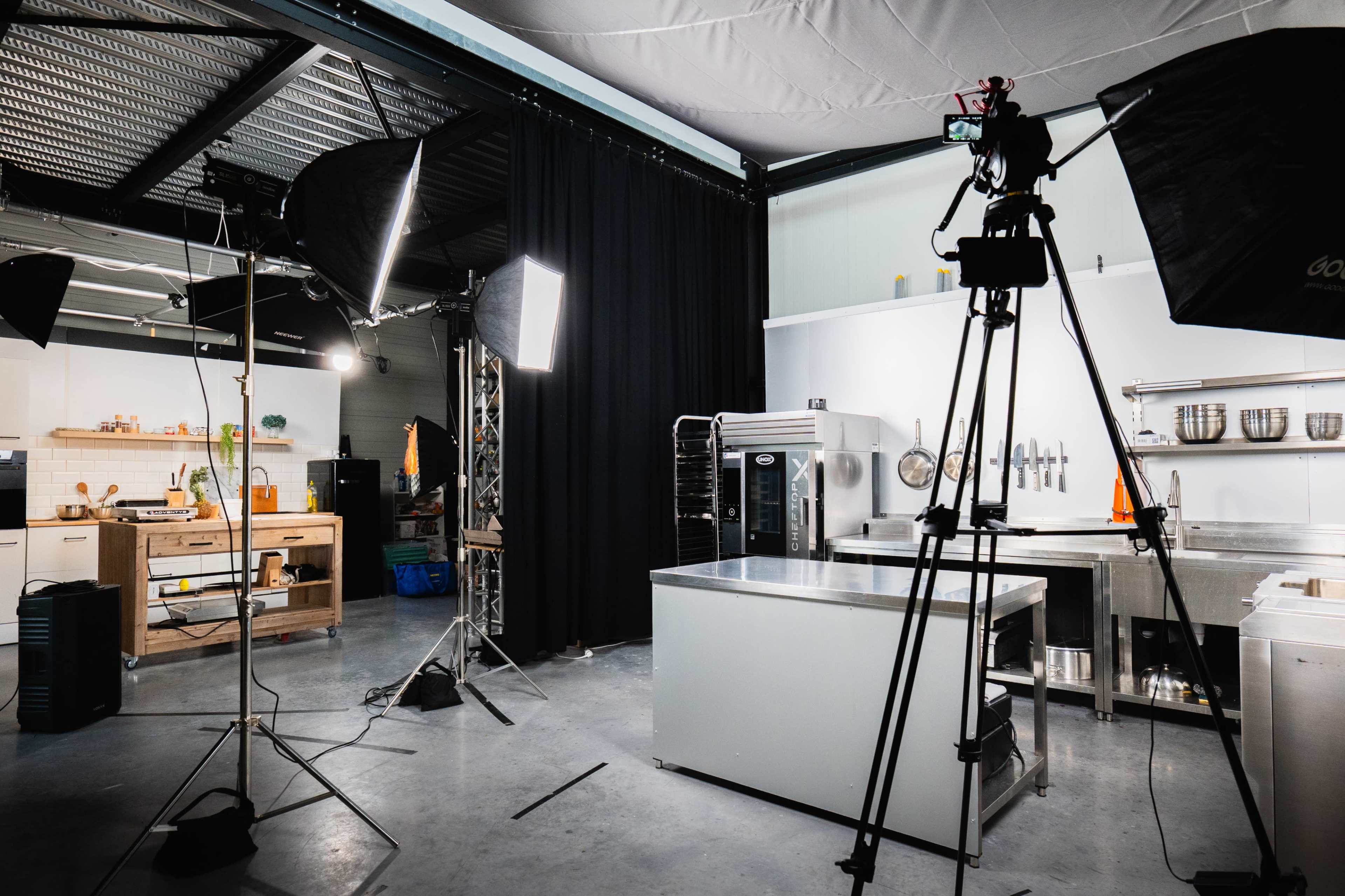 The image shows a well-equipped commercial kitchen set up for a photo or video shoot, with various lighting equipment and a camera on a tripod positioned for recording.