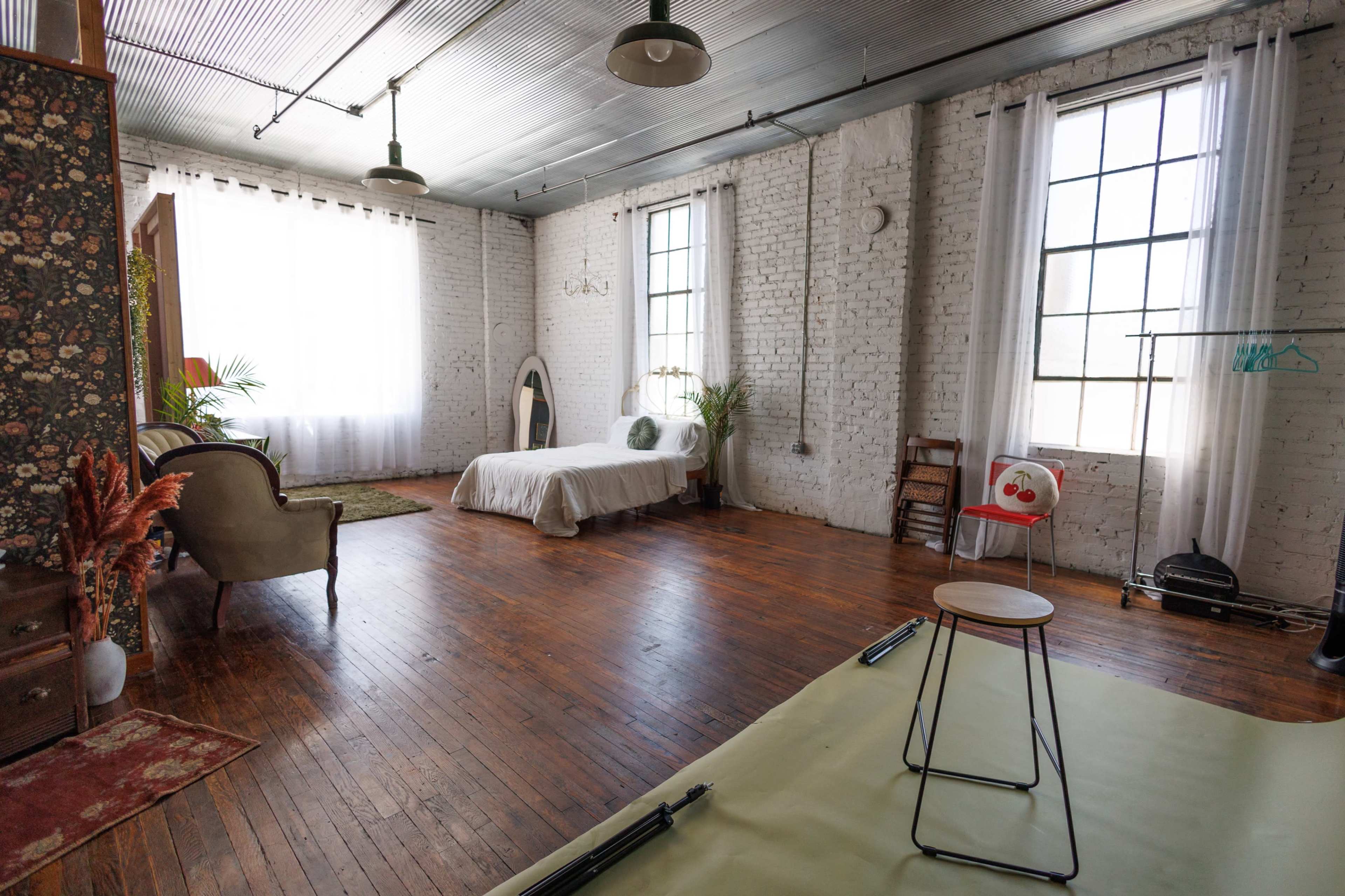 The image shows a spacious room with wooden flooring, large windows dressed in sheer white curtains, a bed with minimal bedding, a vintage armchair, and a stool.