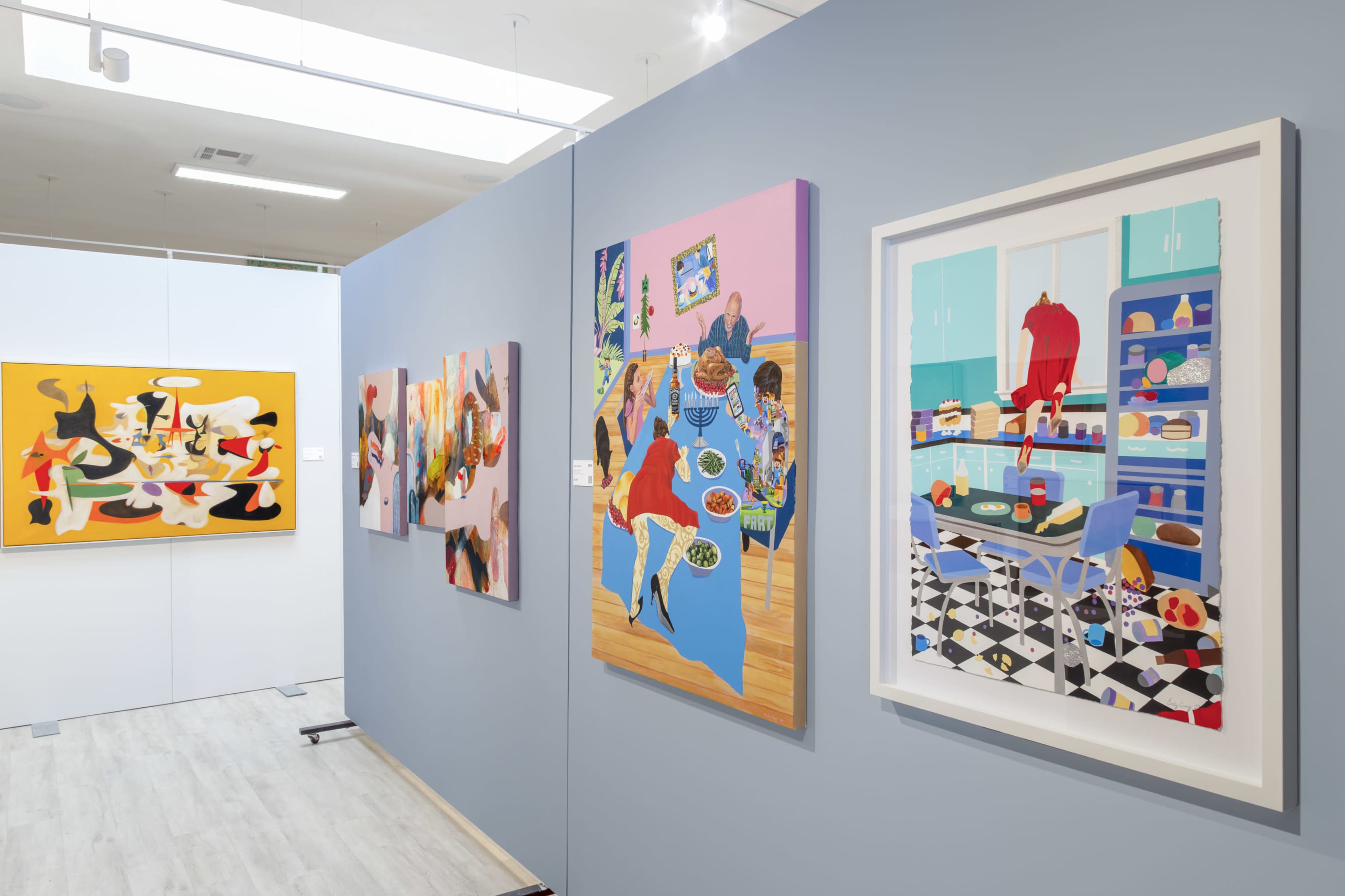The image shows a gallery with colorful, contemporary artwork displayed on light blue walls, featuring various styles and scenes, including people interacting and kitchen settings.