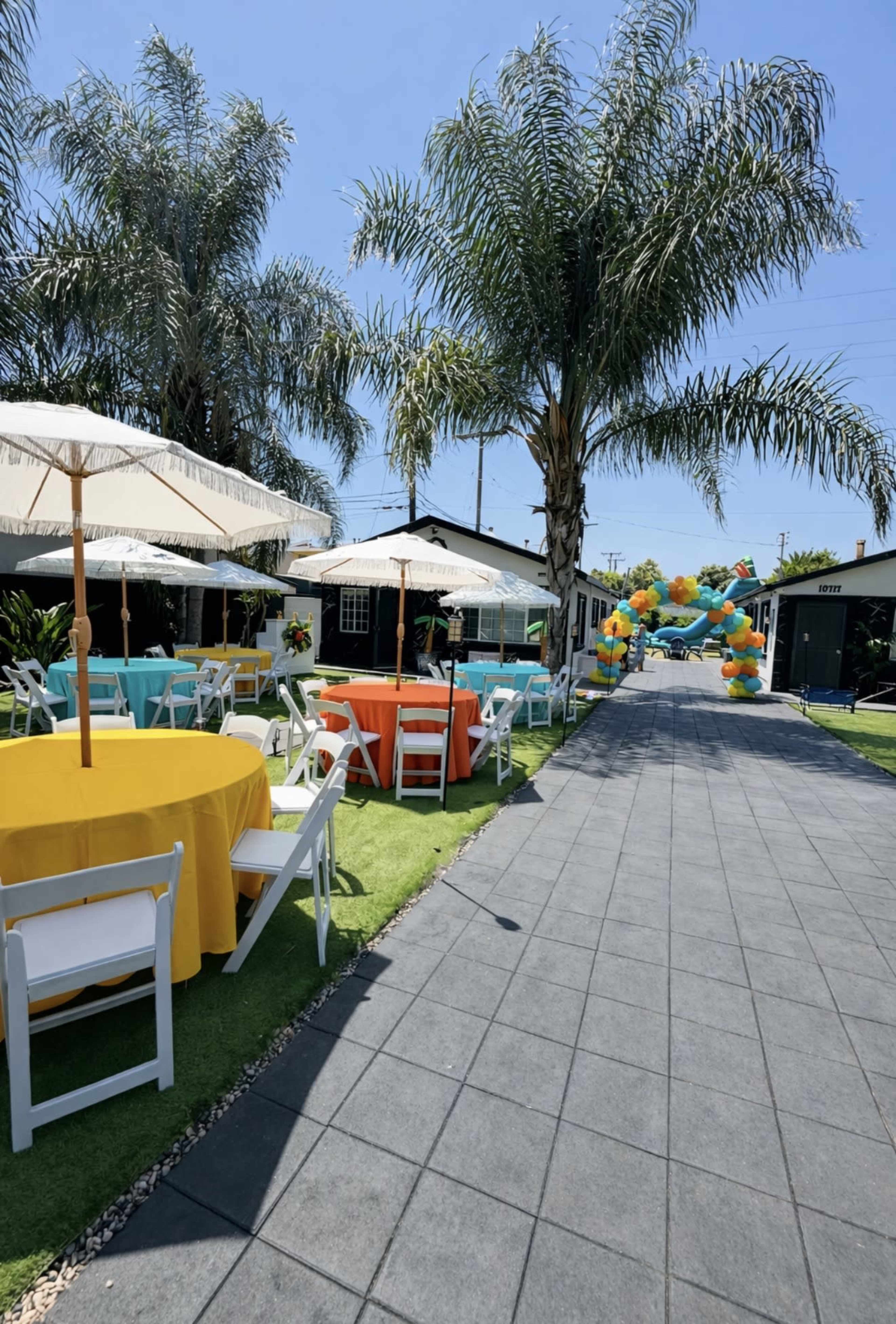 The image shows a party setup with colorful tables under umbrellas, flanked by palm trees and a pathway leading to a balloon arch in the background.