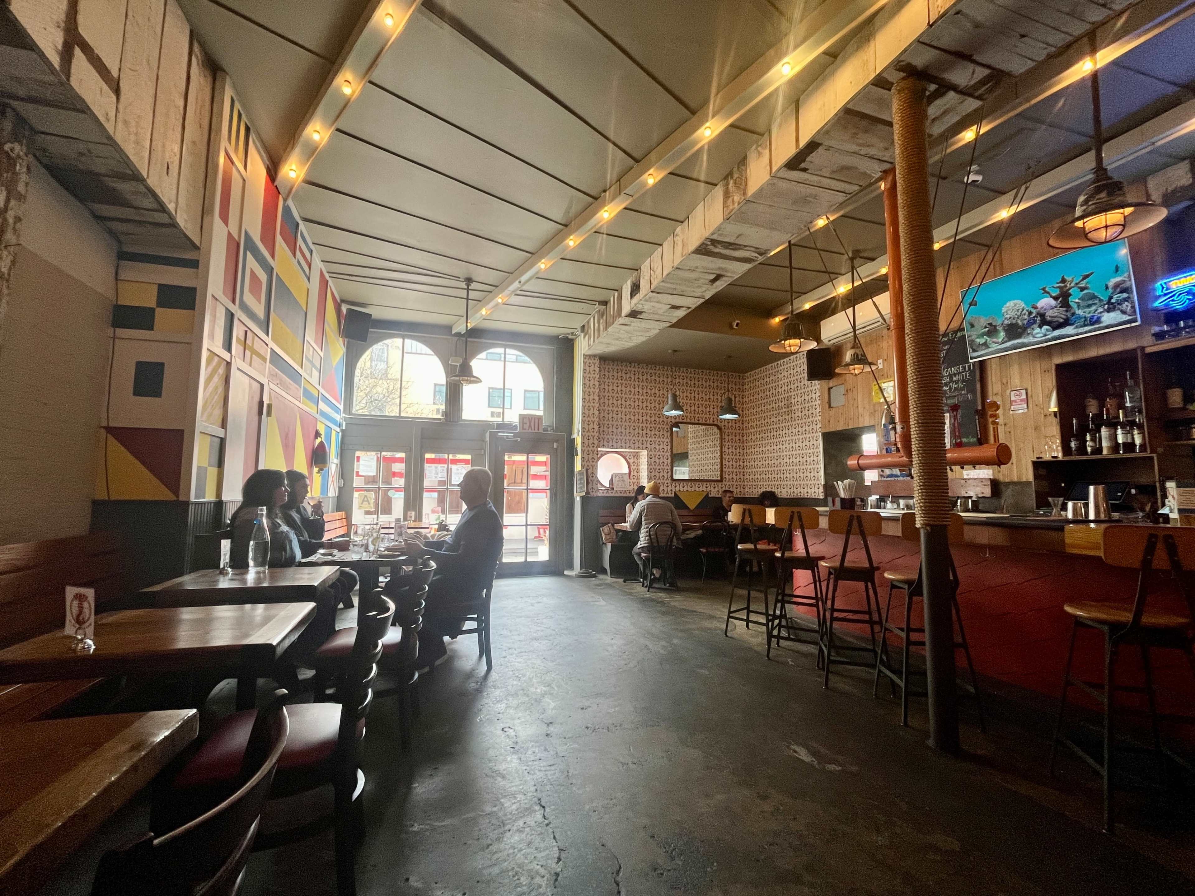 The interior of a restaurant features colorful walls, wooden tables, and patrons seated at tables and the bar, with large windows allowing natural light to fill the space.