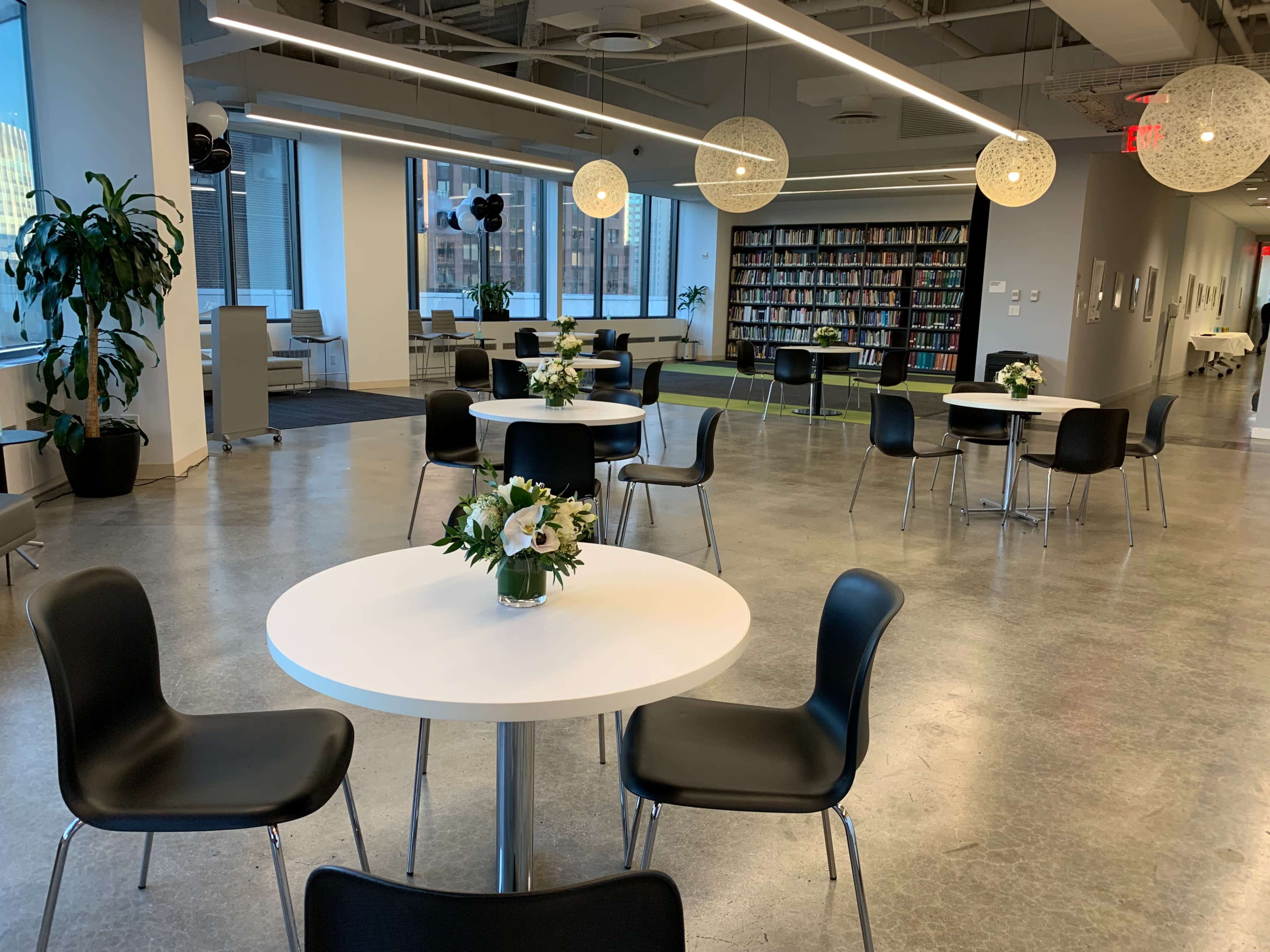 A modern, well-lit open space with round tables, black chairs, and a bookshelf in the background, adorned with floral centerpieces.