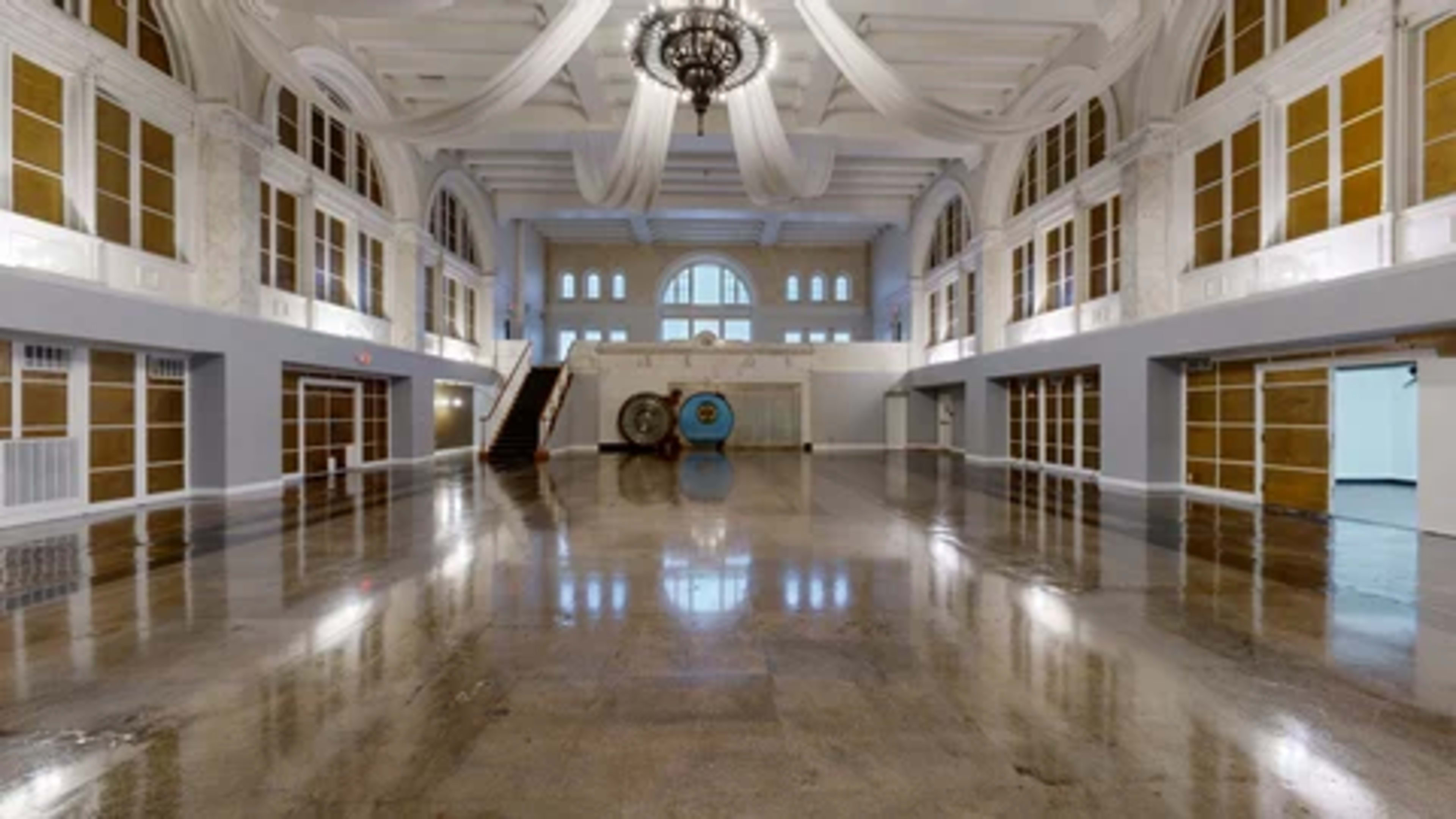 The image shows a spacious, empty hall with high ceilings, large windows, and polished floors reflecting the overhead chandelier and architectural details.