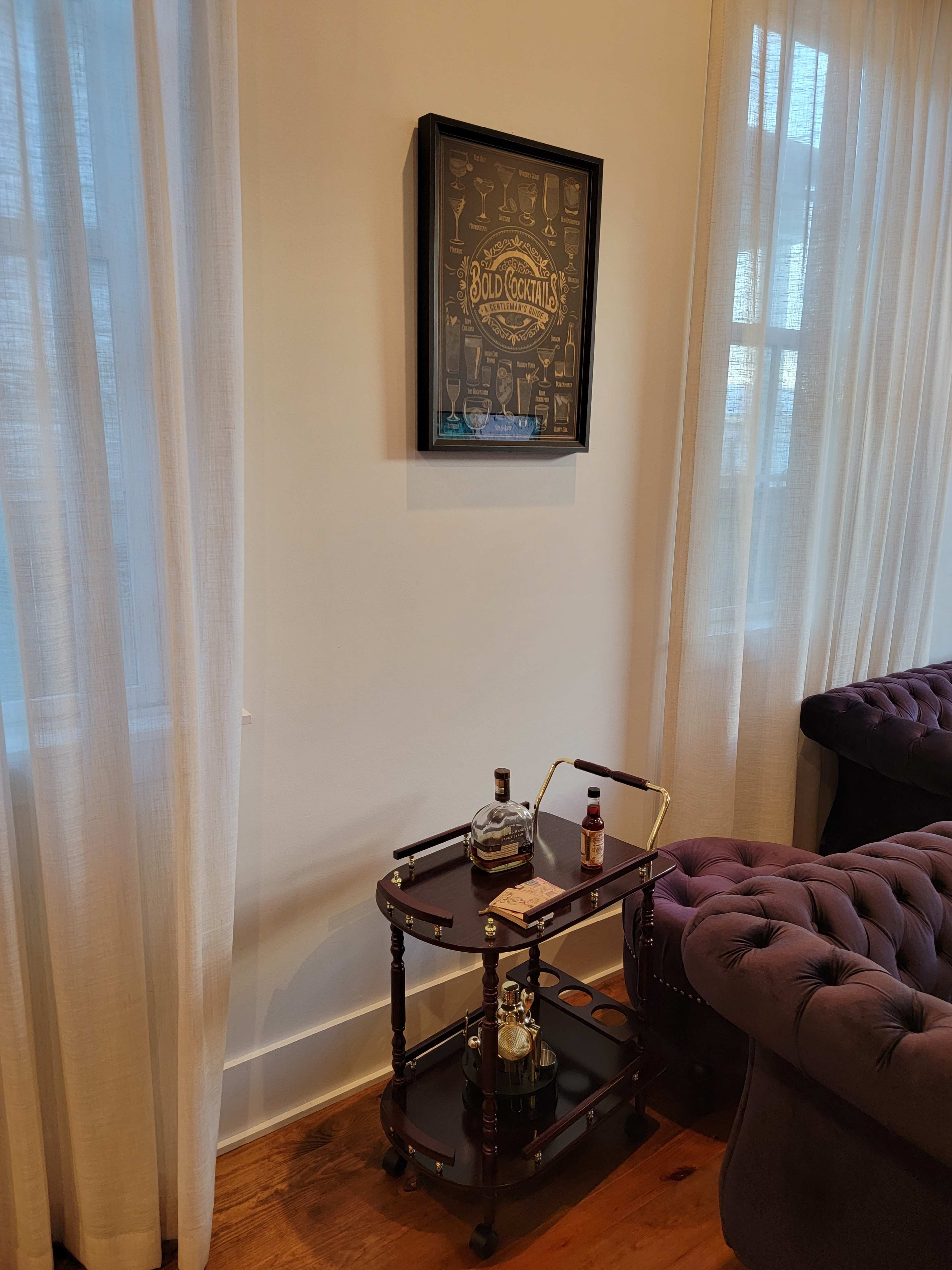 A vintage bar cart with a selection of spirits is situated next to a framed cocktail poster on a white wall, illuminated by natural light through sheer curtains.