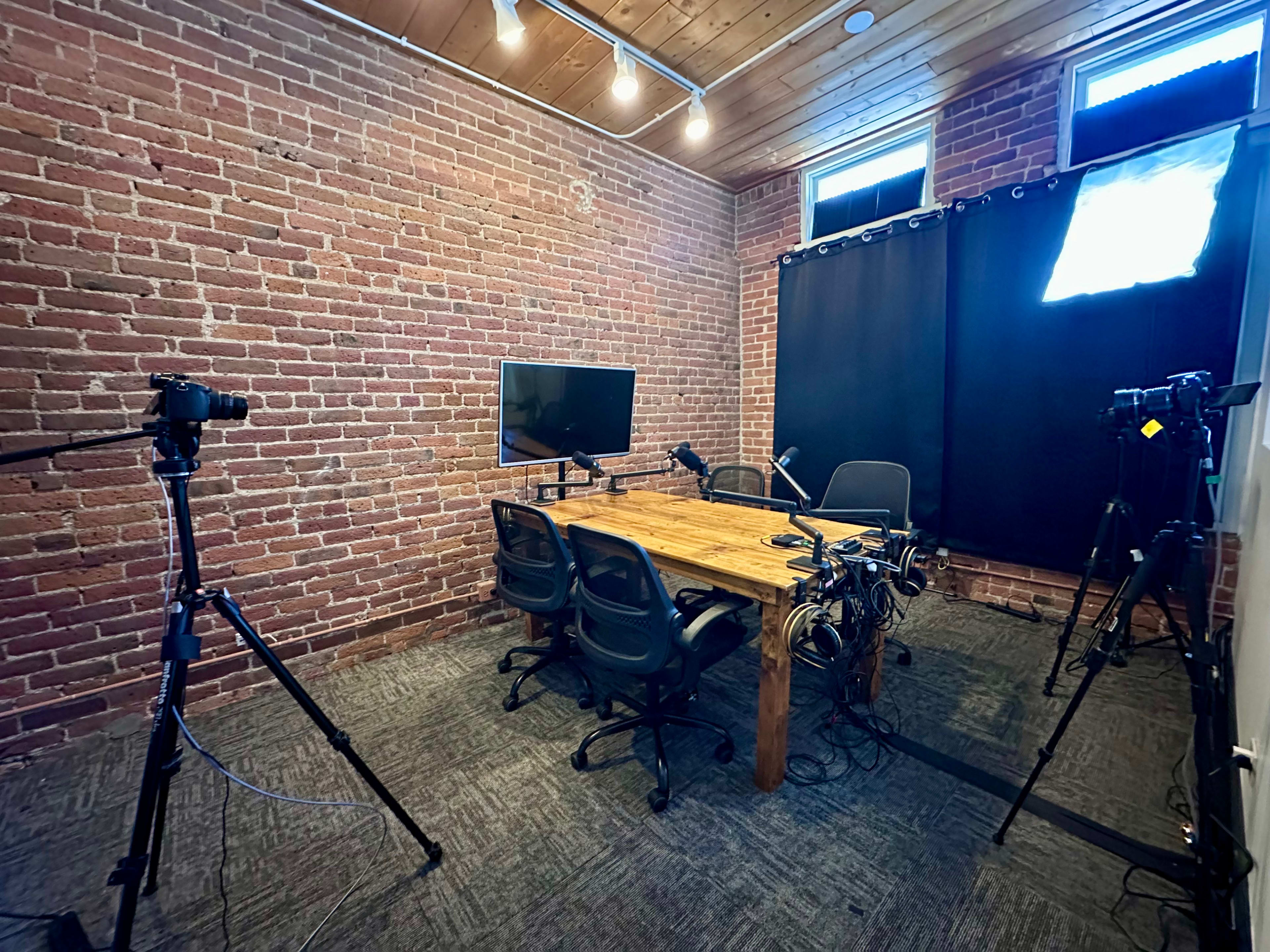 The image shows a small studio with exposed brick walls, a wooden table surrounded by chairs, and multiple cameras set up, including a monitor and lighting equipment.