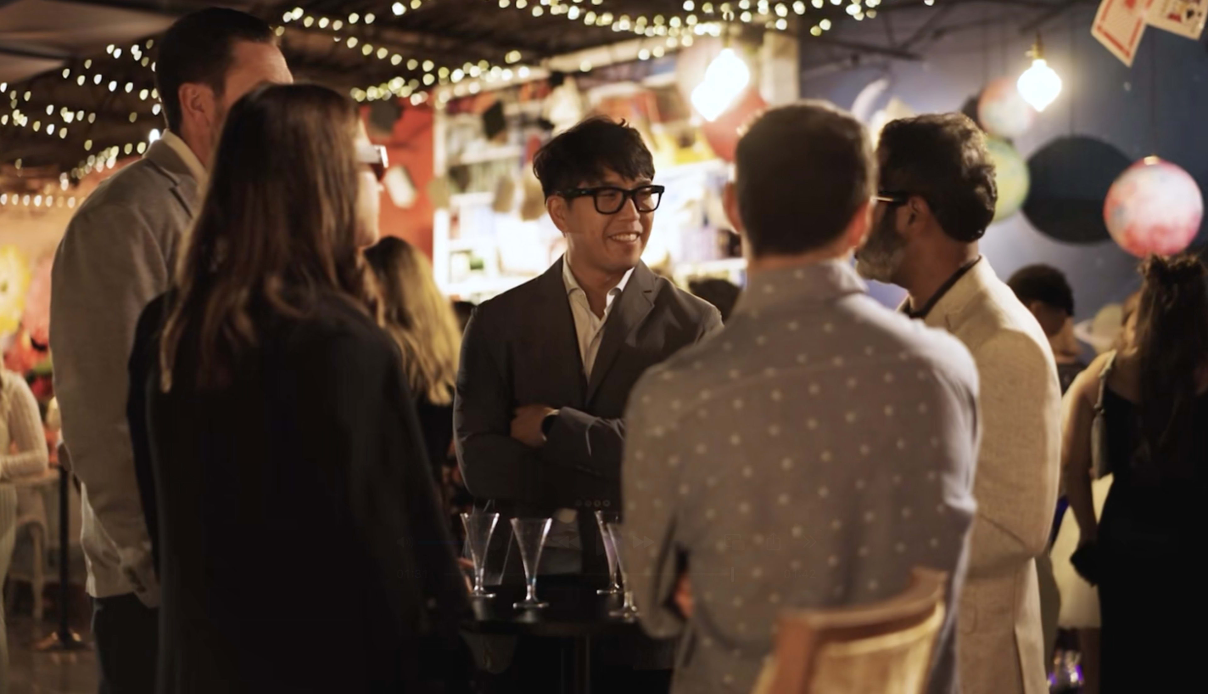 A group of five people, dressed in formal and semi-formal attire, engage in conversation at a lively social event decorated with string lights in the background.