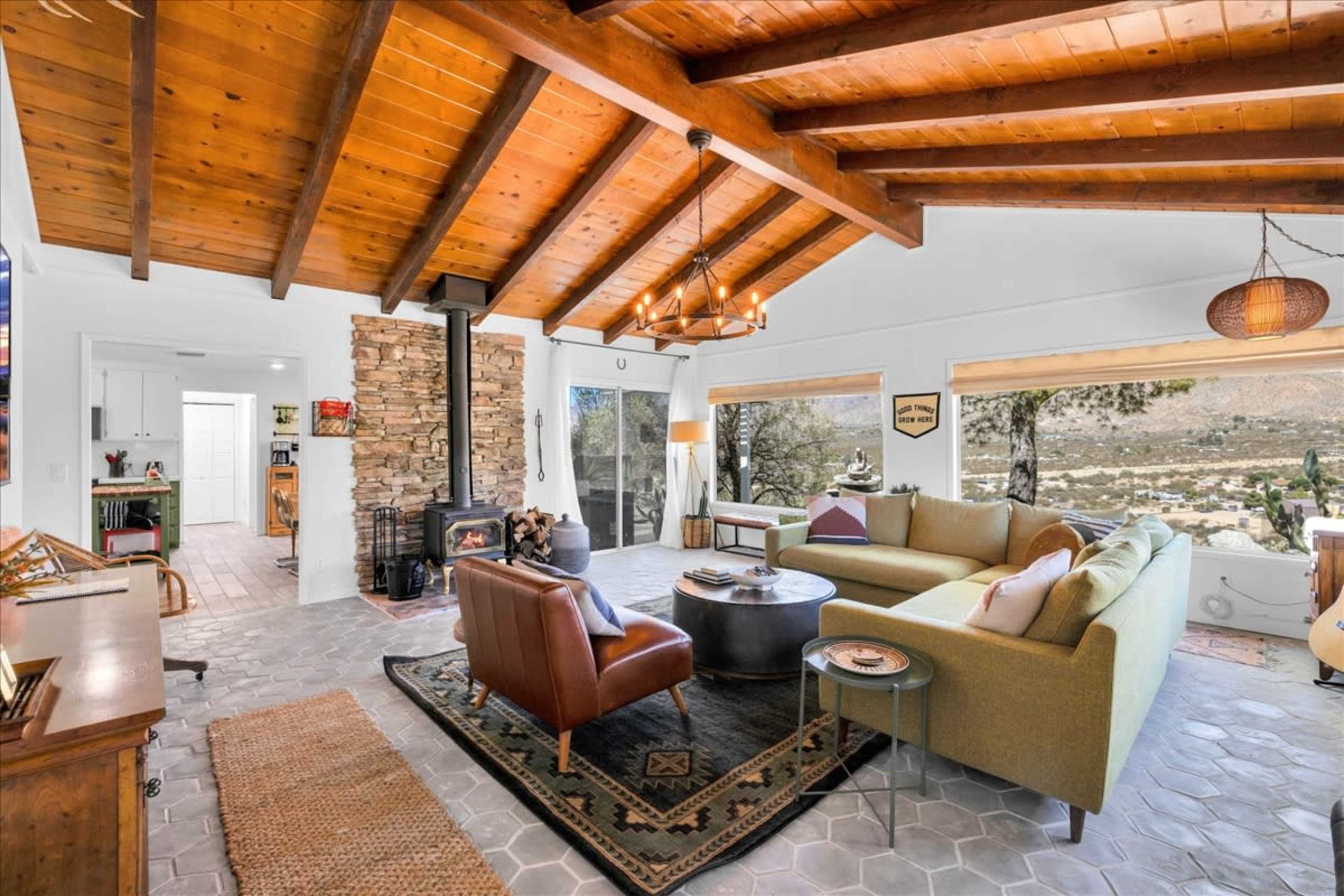 The image shows a cozy living room with a stone fireplace, wooden beams on the ceiling, and a mix of modern and vintage furniture, set against a backdrop of a scenic view through large windows.