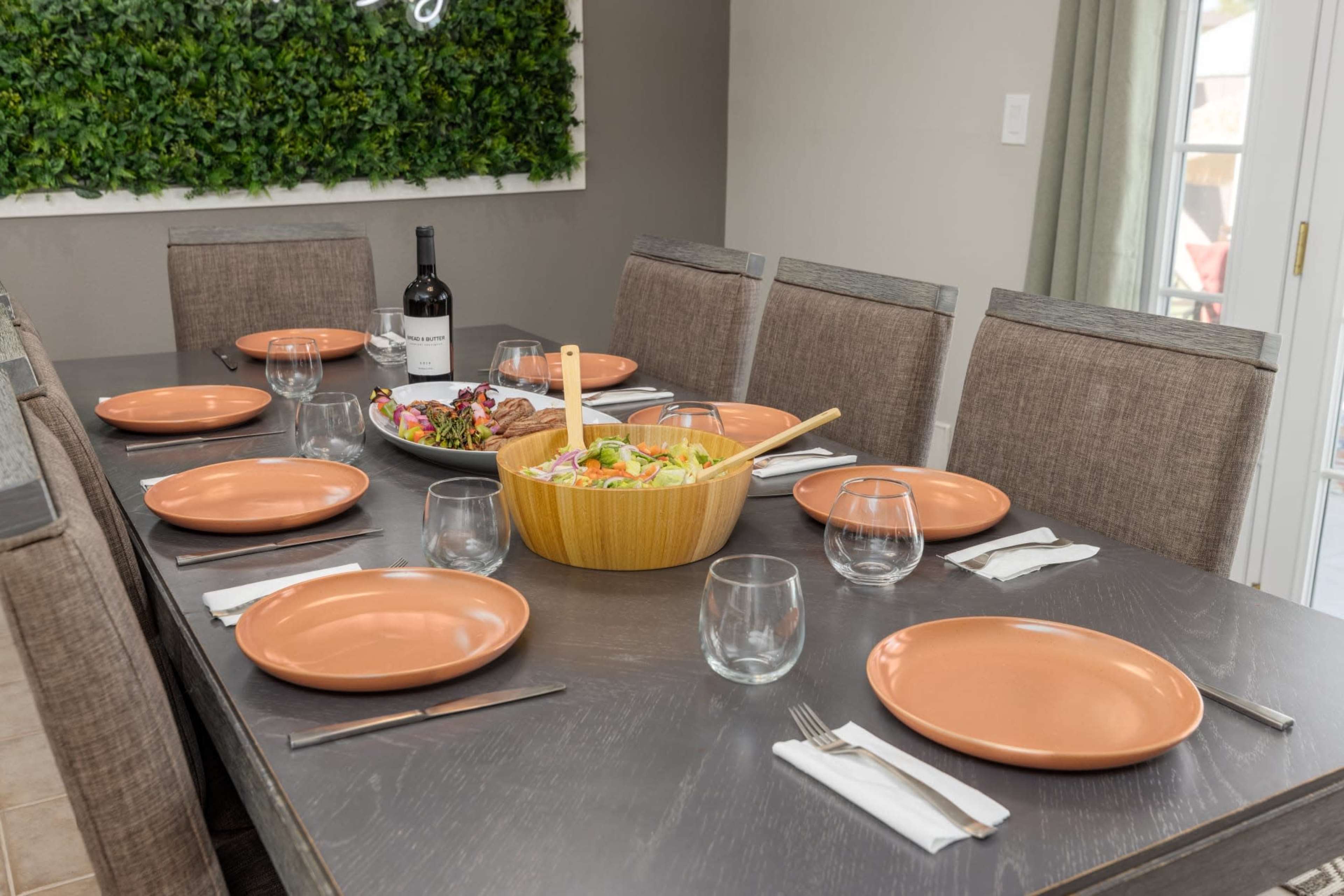 A dining table is set with orange plates, glassware, and a wooden bowl of salad, accompanied by a bottle of wine and a colorful platter of food.