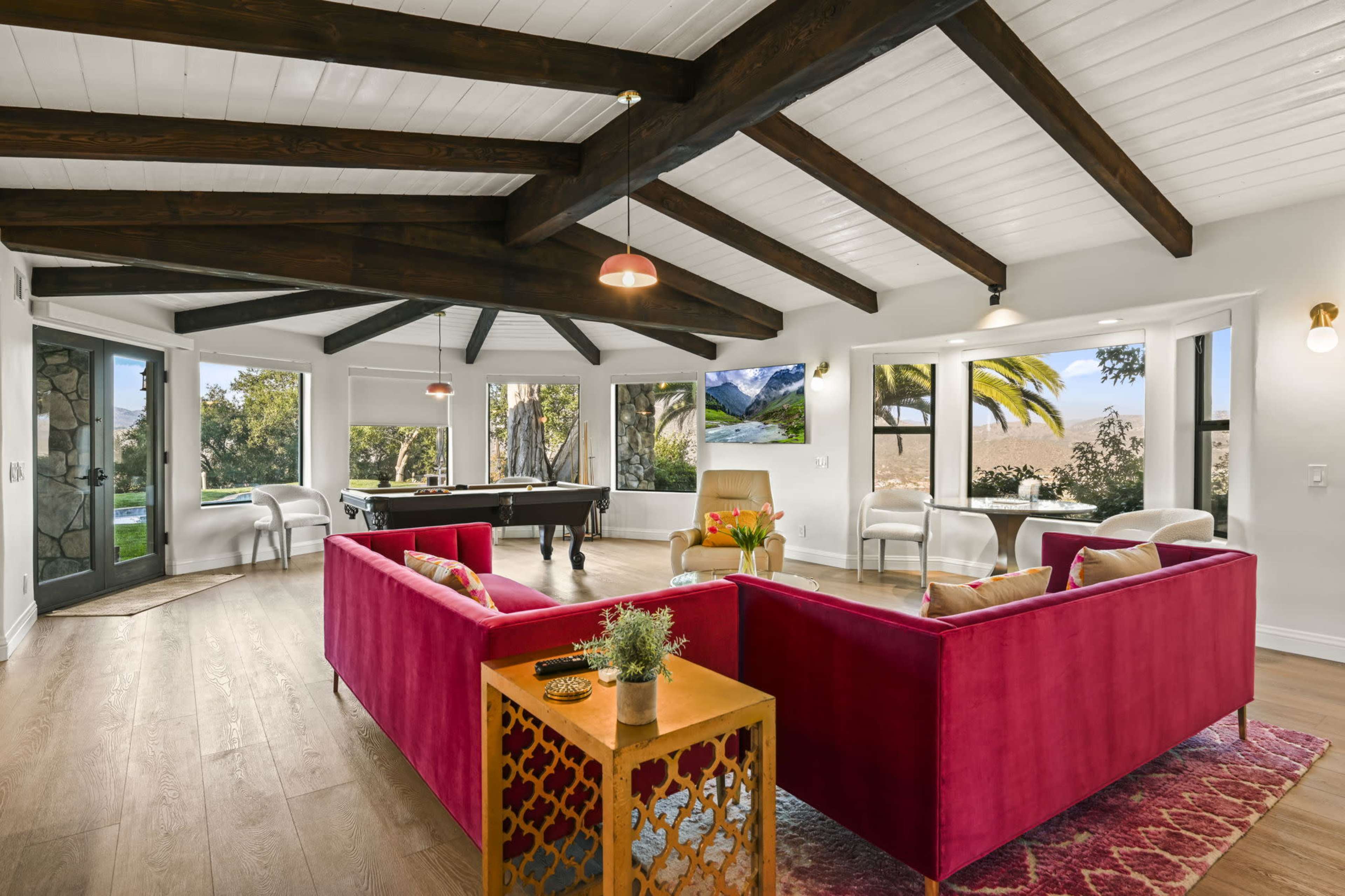 The image shows a spacious living area featuring a pink sectional sofa, a wooden table, and large windows that provide natural light and a view of the outdoors.