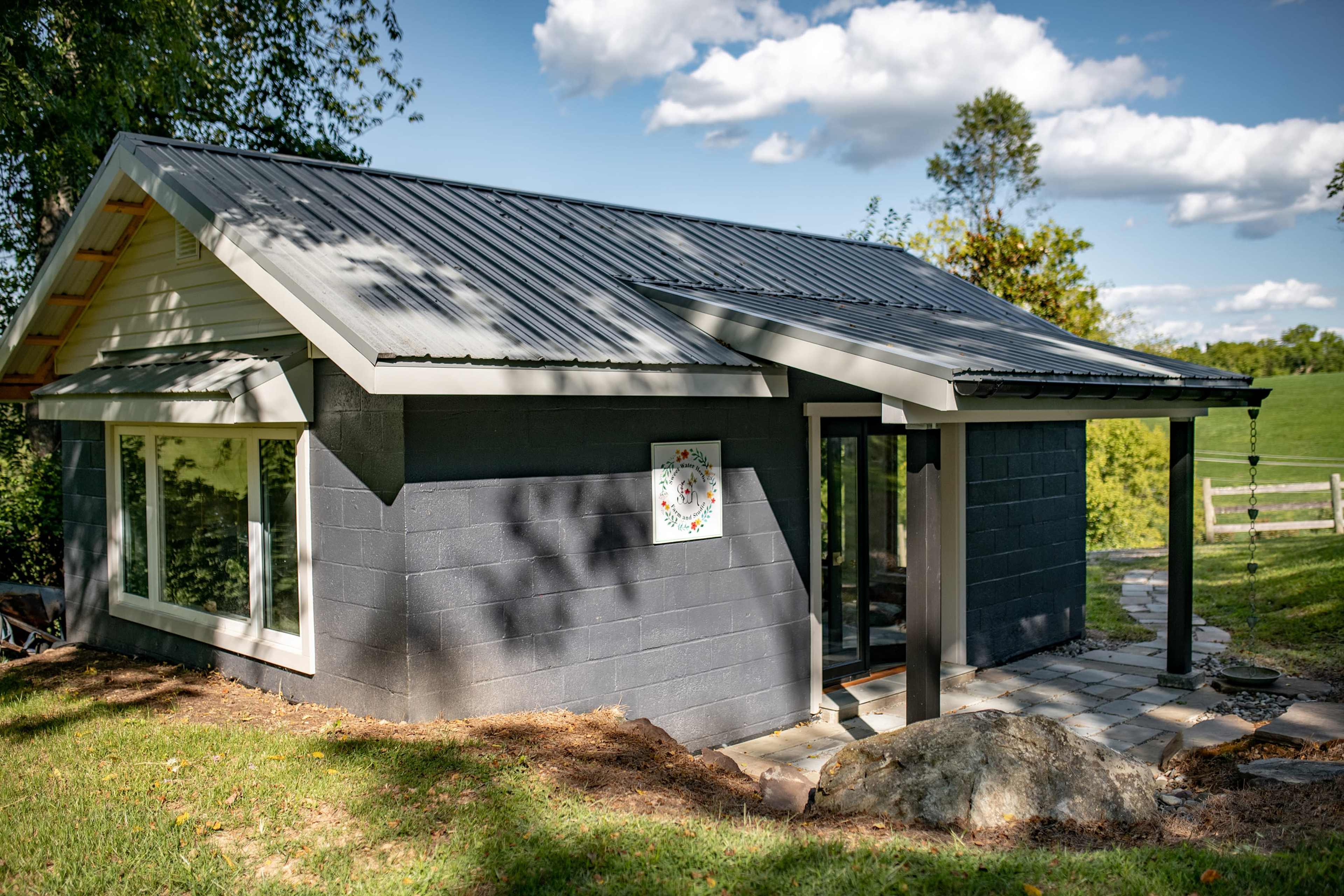 A modern building with a gray exterior and a metal roof is surrounded by greenery and features large windows.