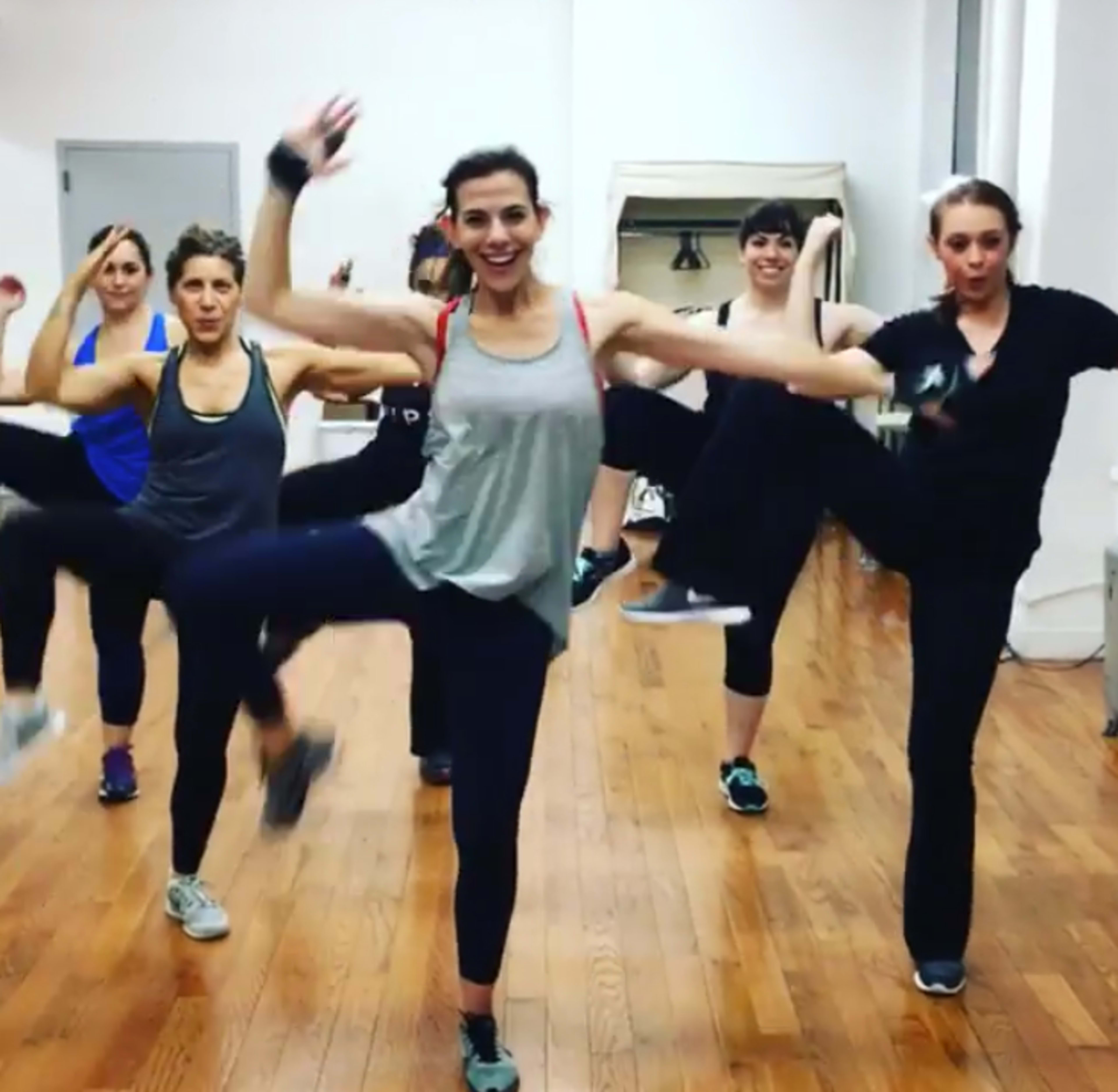 A group of six women are participating in a dance or exercise class, raising their legs in a synchronized manner in a well-lit studio with wooden flooring.