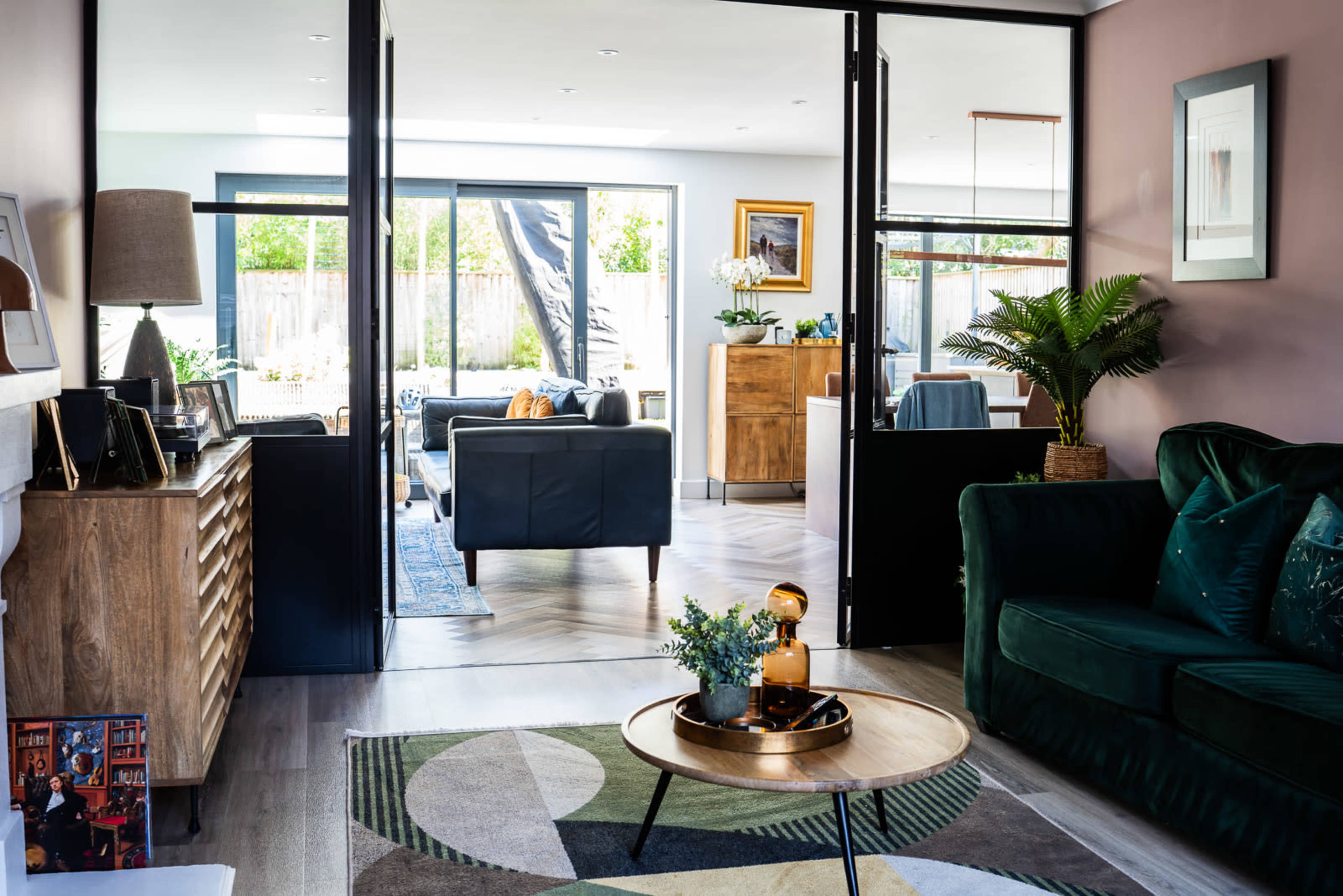 The image shows a modern living space divided by glass doors, featuring a green sofa, a wooden sideboard, and a view into a bright, well-furnished room with outdoor access.