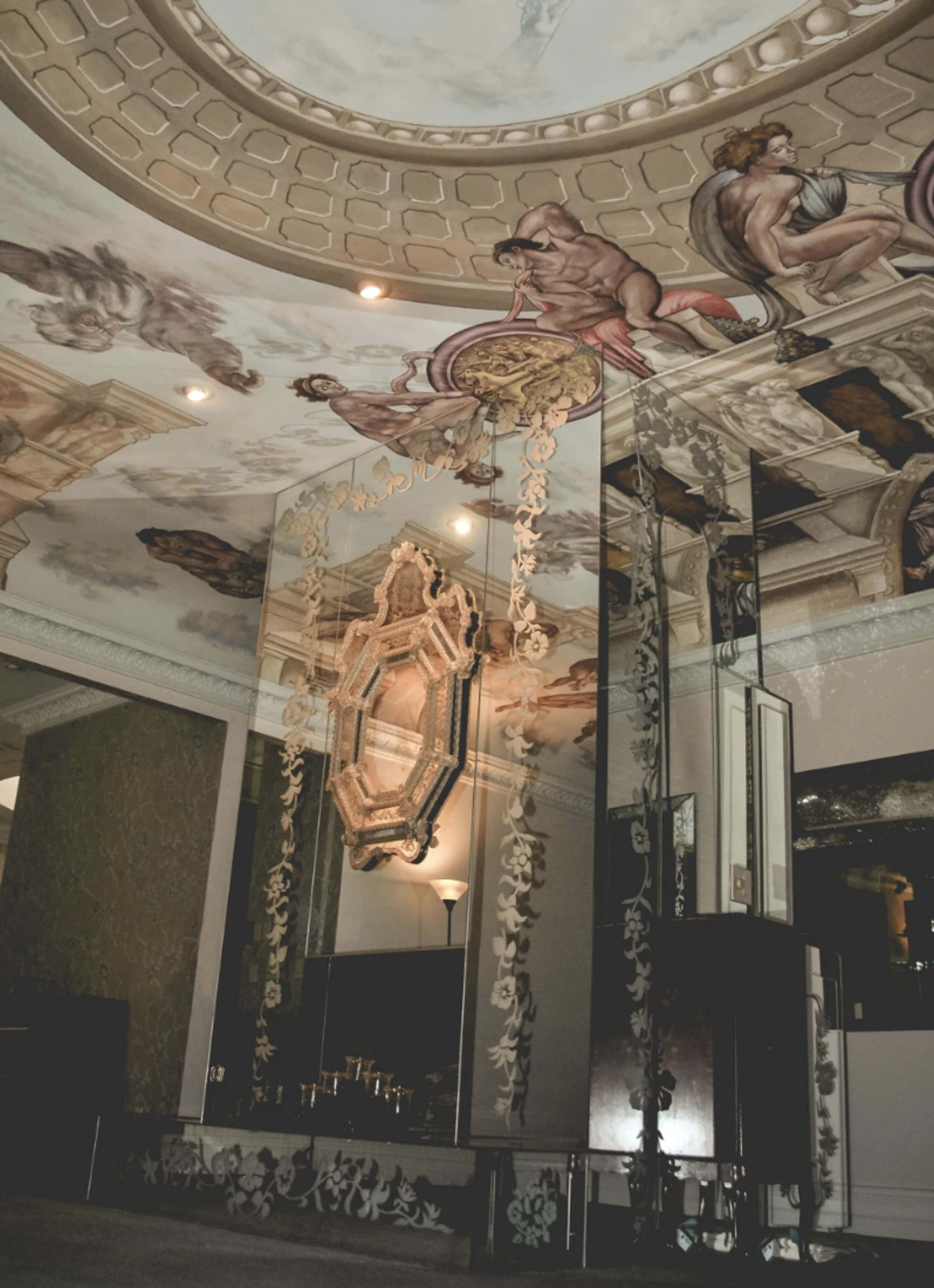 An elegant interior space featuring ornate ceiling frescoes, a large mirror with intricate carvings, and decorative wall elements.