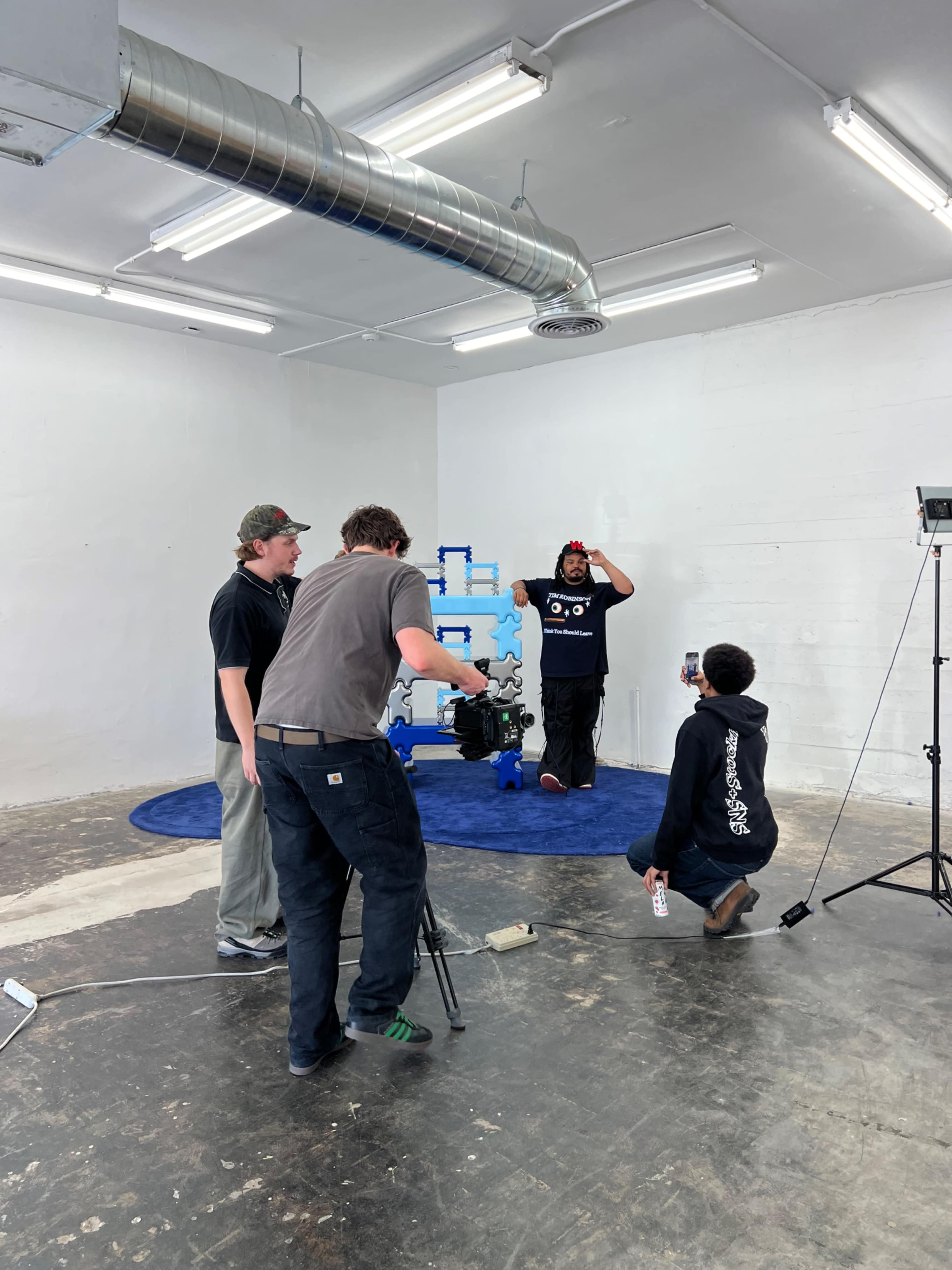 A group of four people is engaged in a video production setup in a minimalistic studio space with white walls and a blue circular rug.
