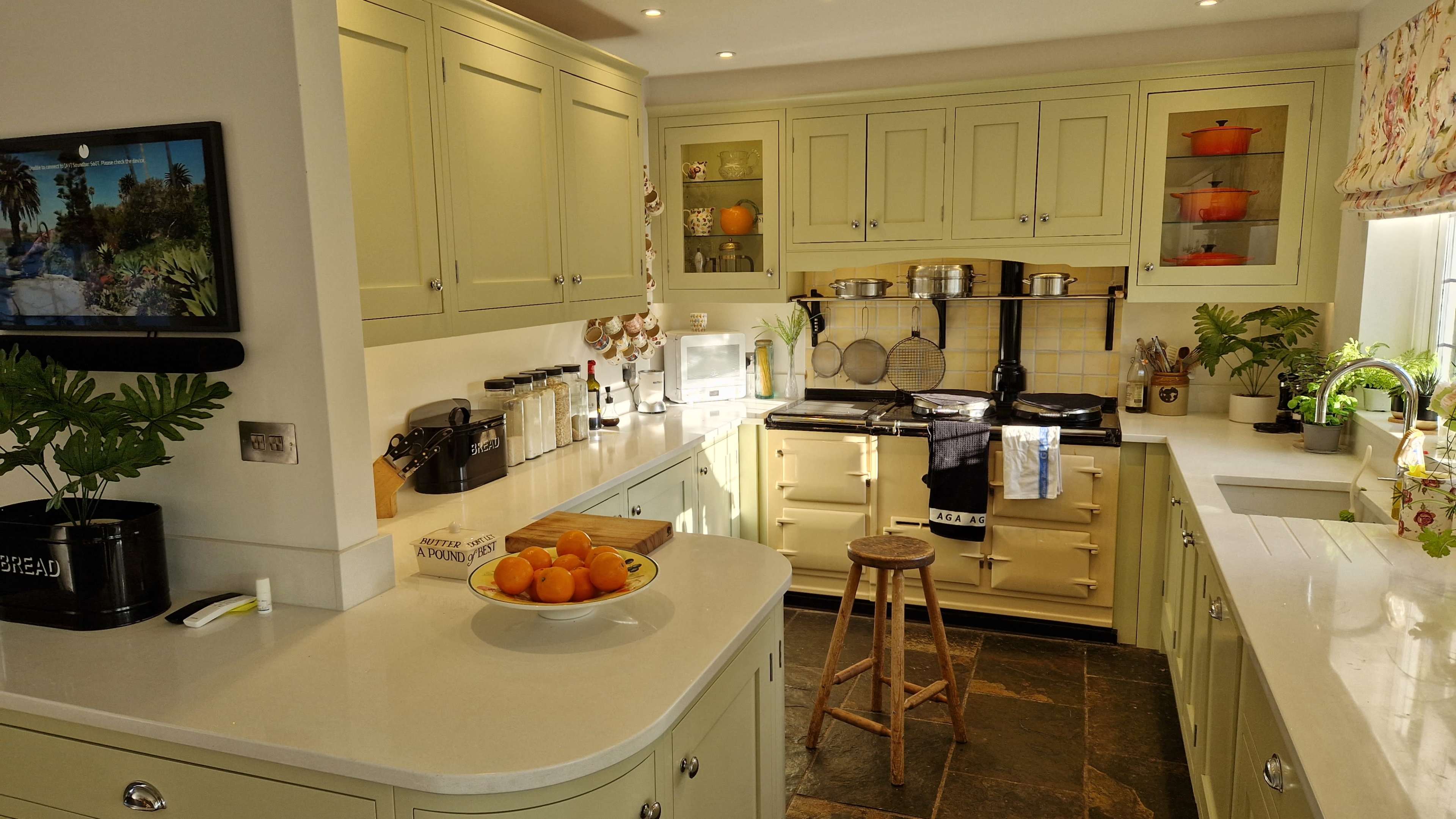 A brightly lit kitchen features green cabinets, a white countertop, an gas stove, and a round wooden stool beside a bowl of oranges.