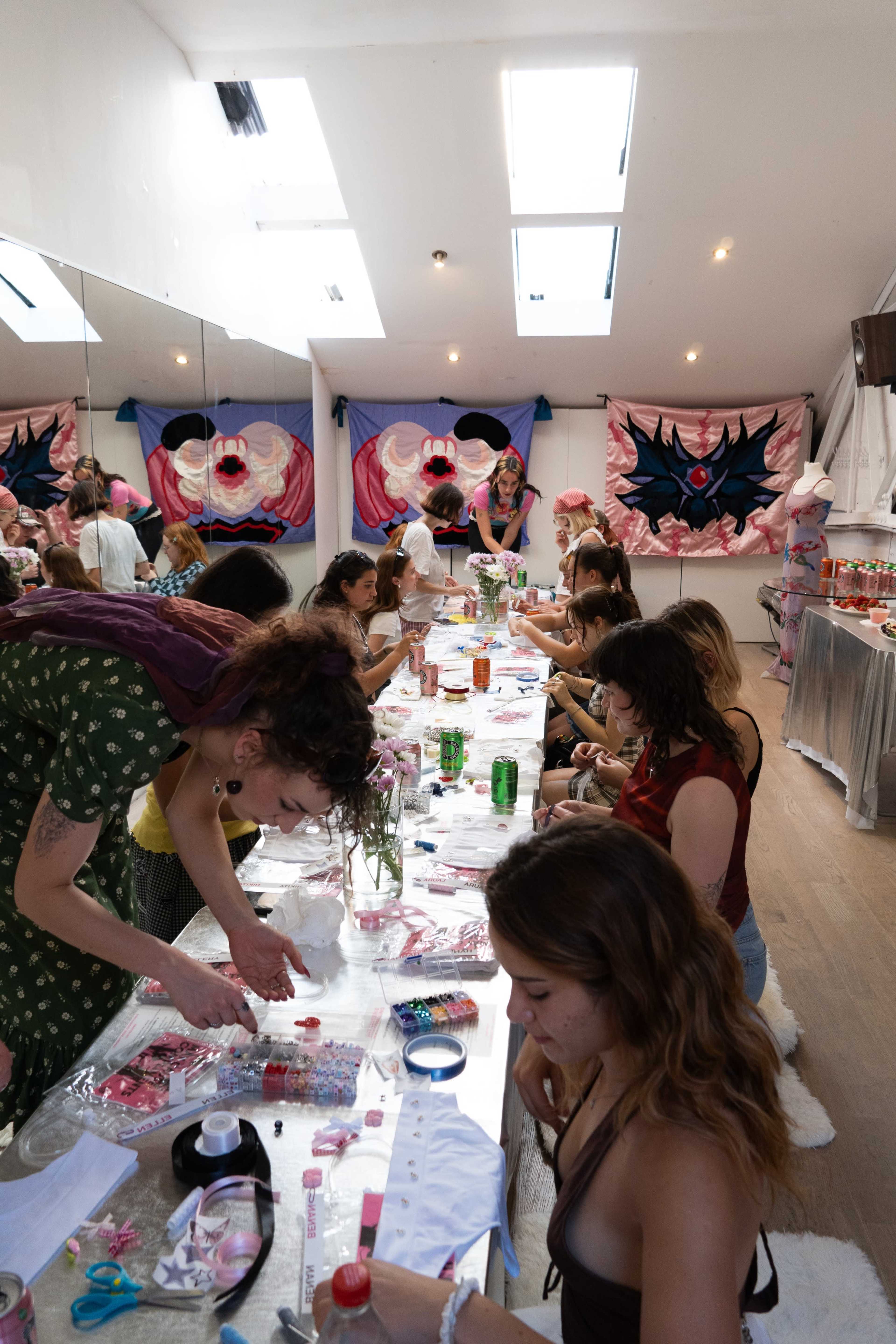 A group of people is engaged in a crafting activity at a long table in a brightly lit room with decorative fabrics on the walls.