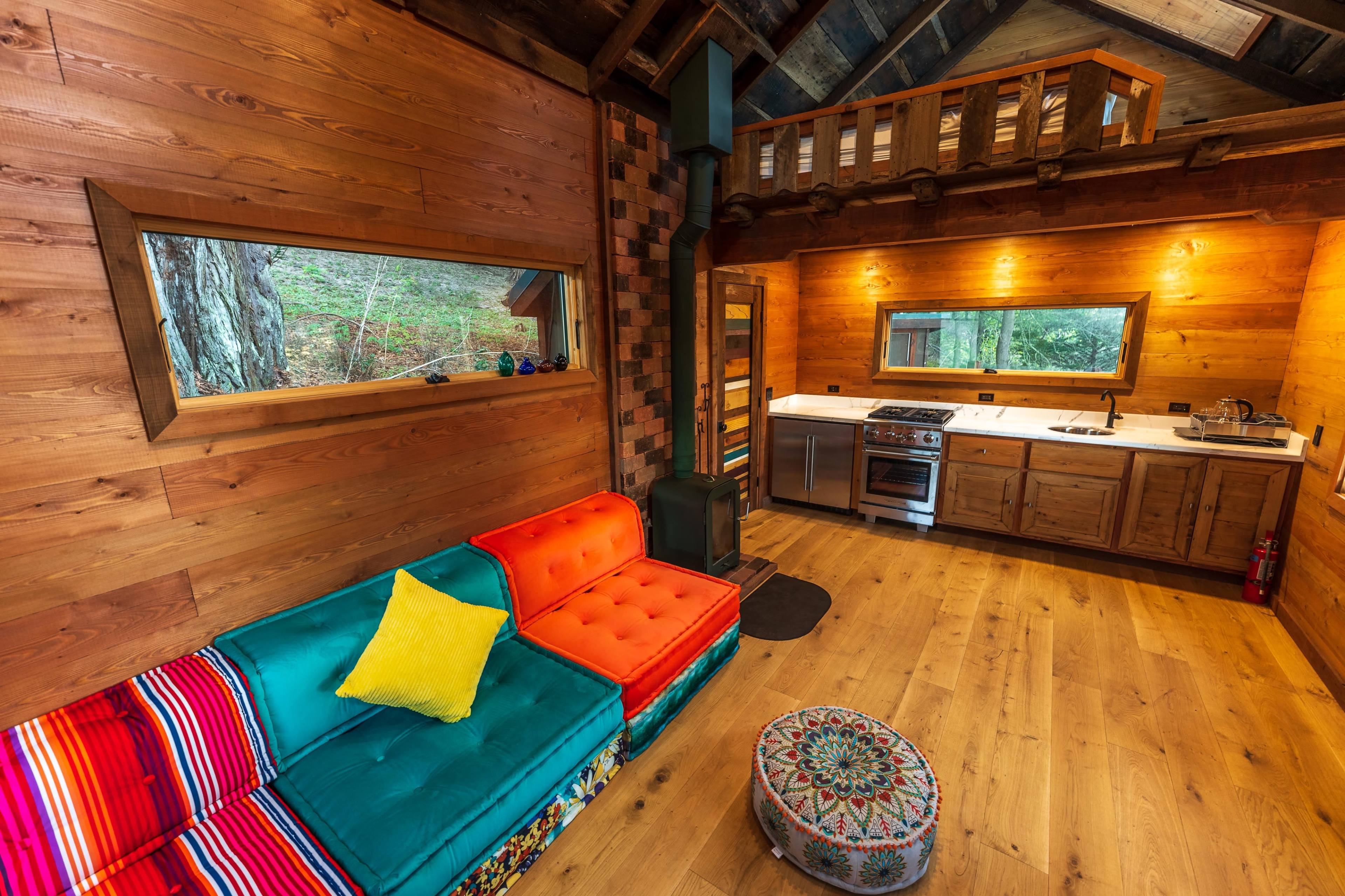 The image shows a wooden interior of a cozy cabin with a colorful sofa, a kitchen area, and a lofted sleeping space.