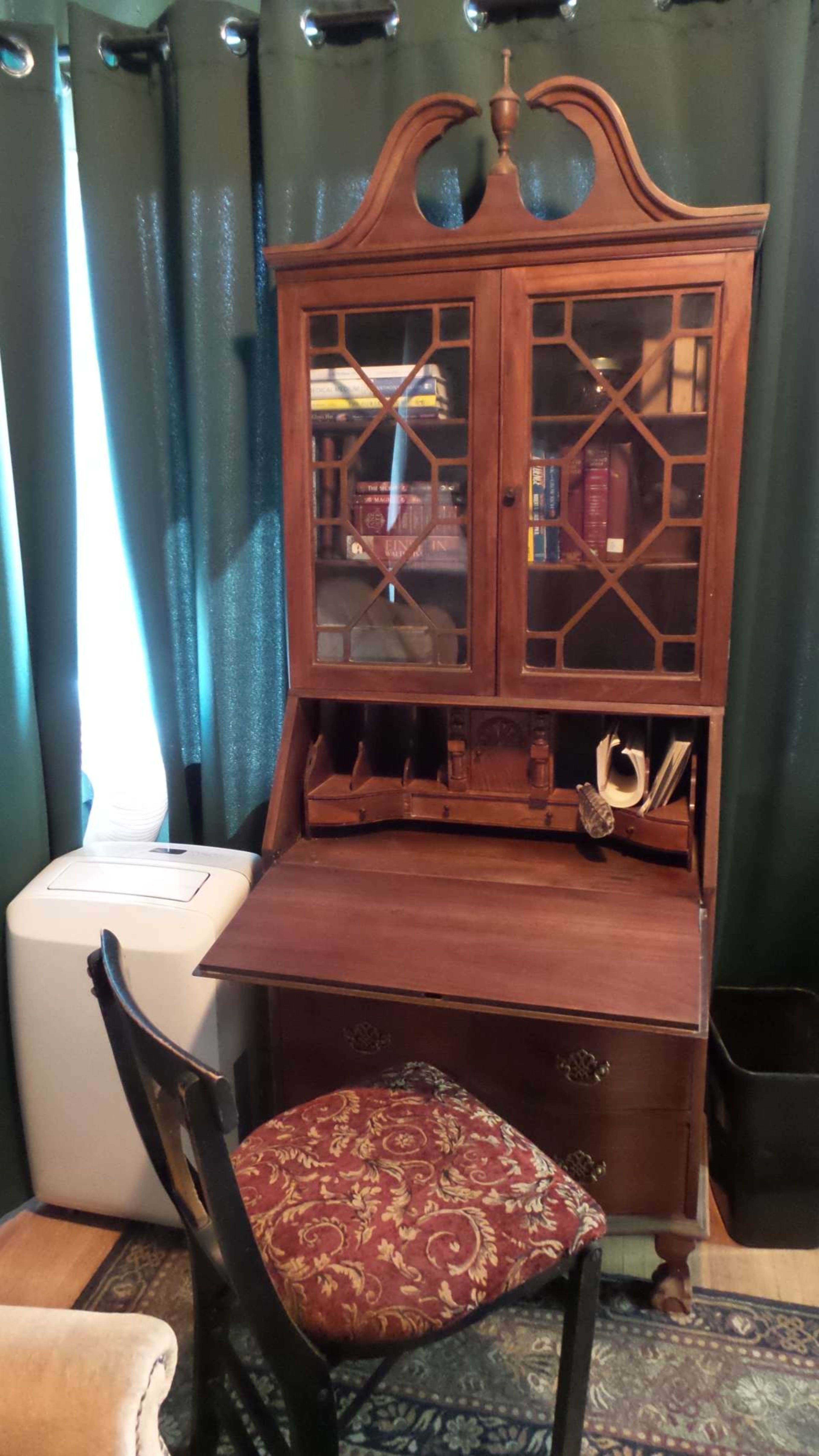 A wooden desk with a hutch and glass-paneled doors sits against a backdrop of dark green curtains, accompanied by a patterned chair.