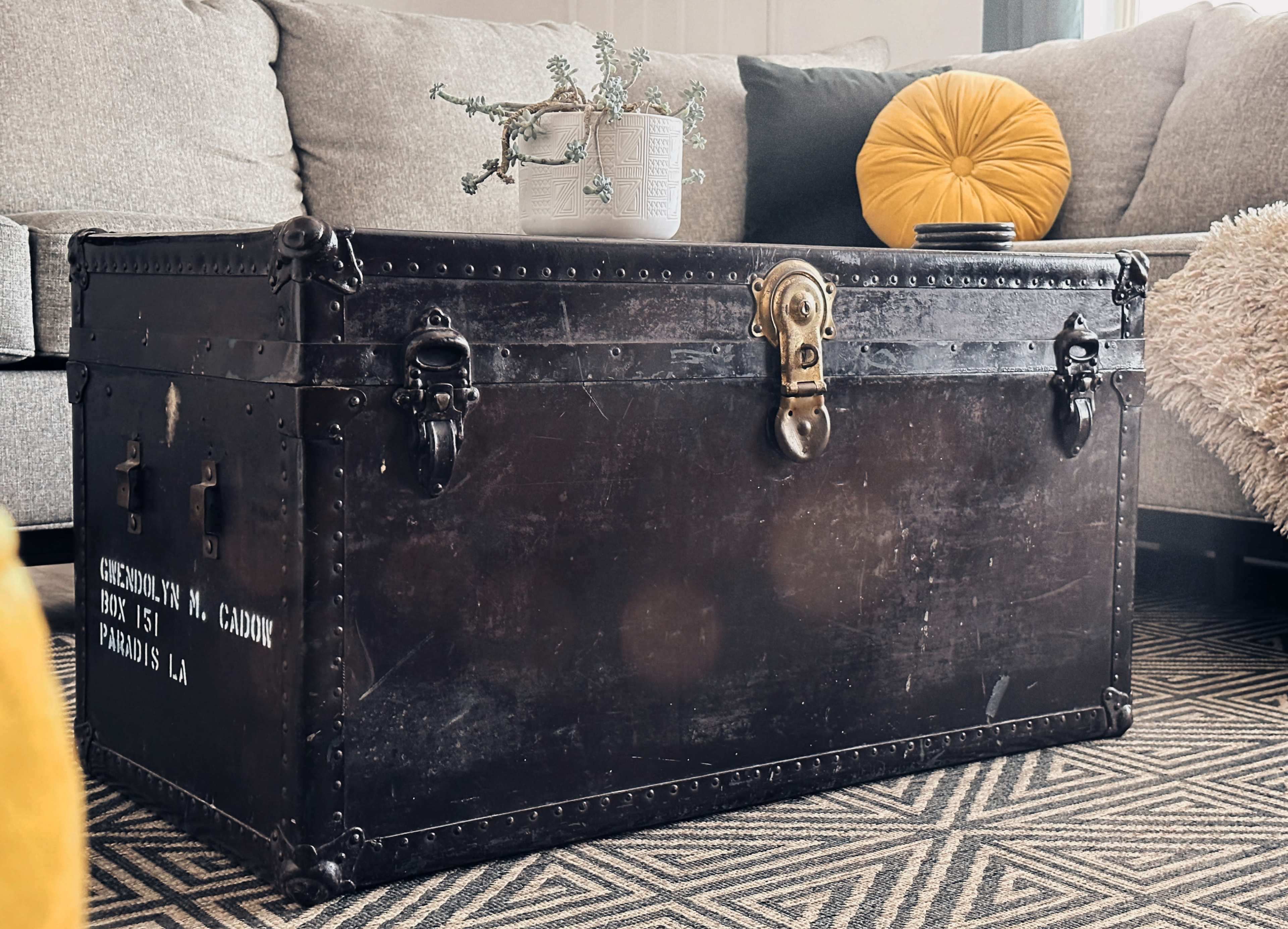 An antique trunk with metal accents sits on a patterned rug in a living room, accompanied by a potted plant and a yellow cushion.