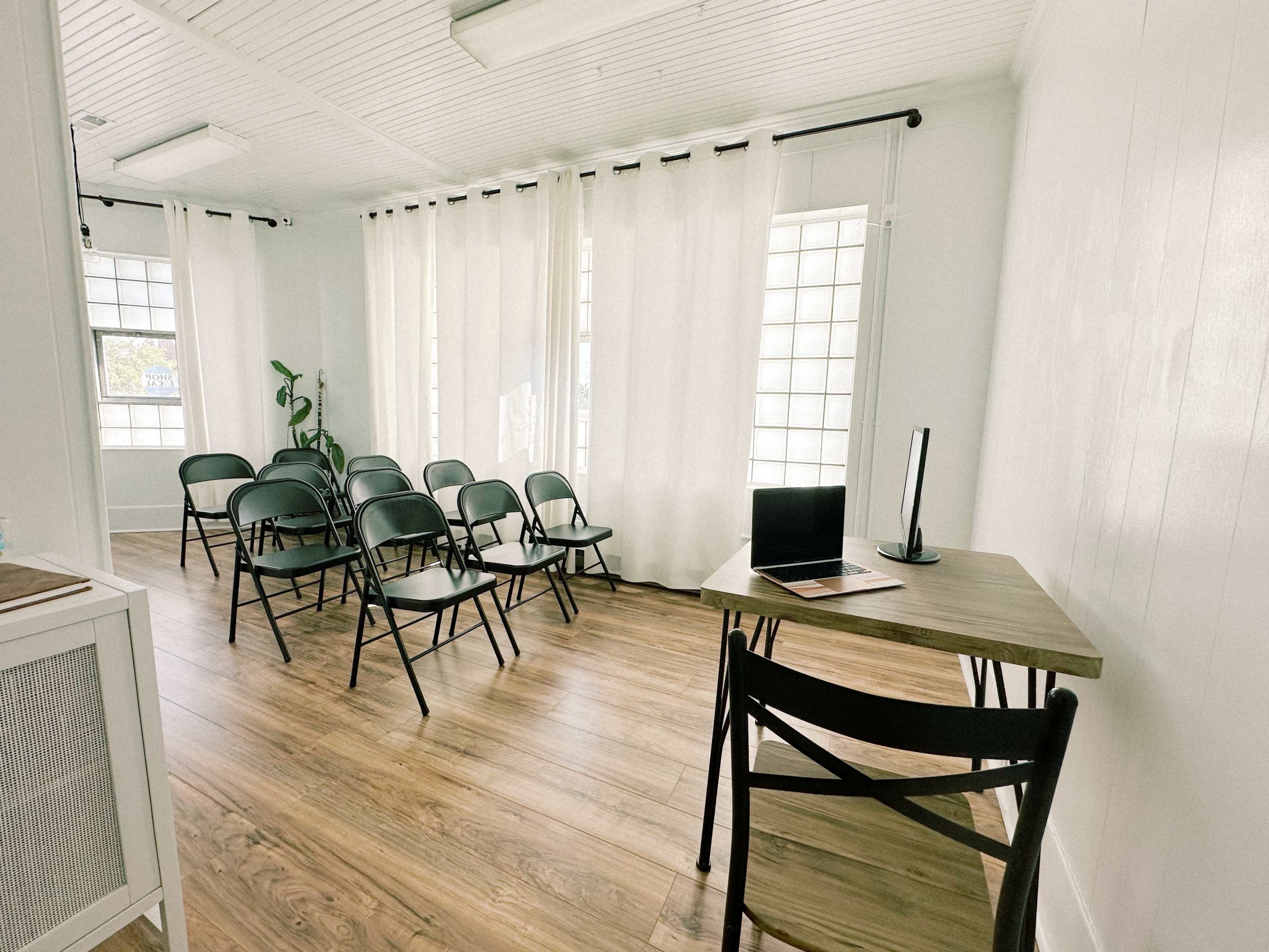 A minimalist meeting room with several black chairs arranged in a semi-circle around a desk with a laptop.