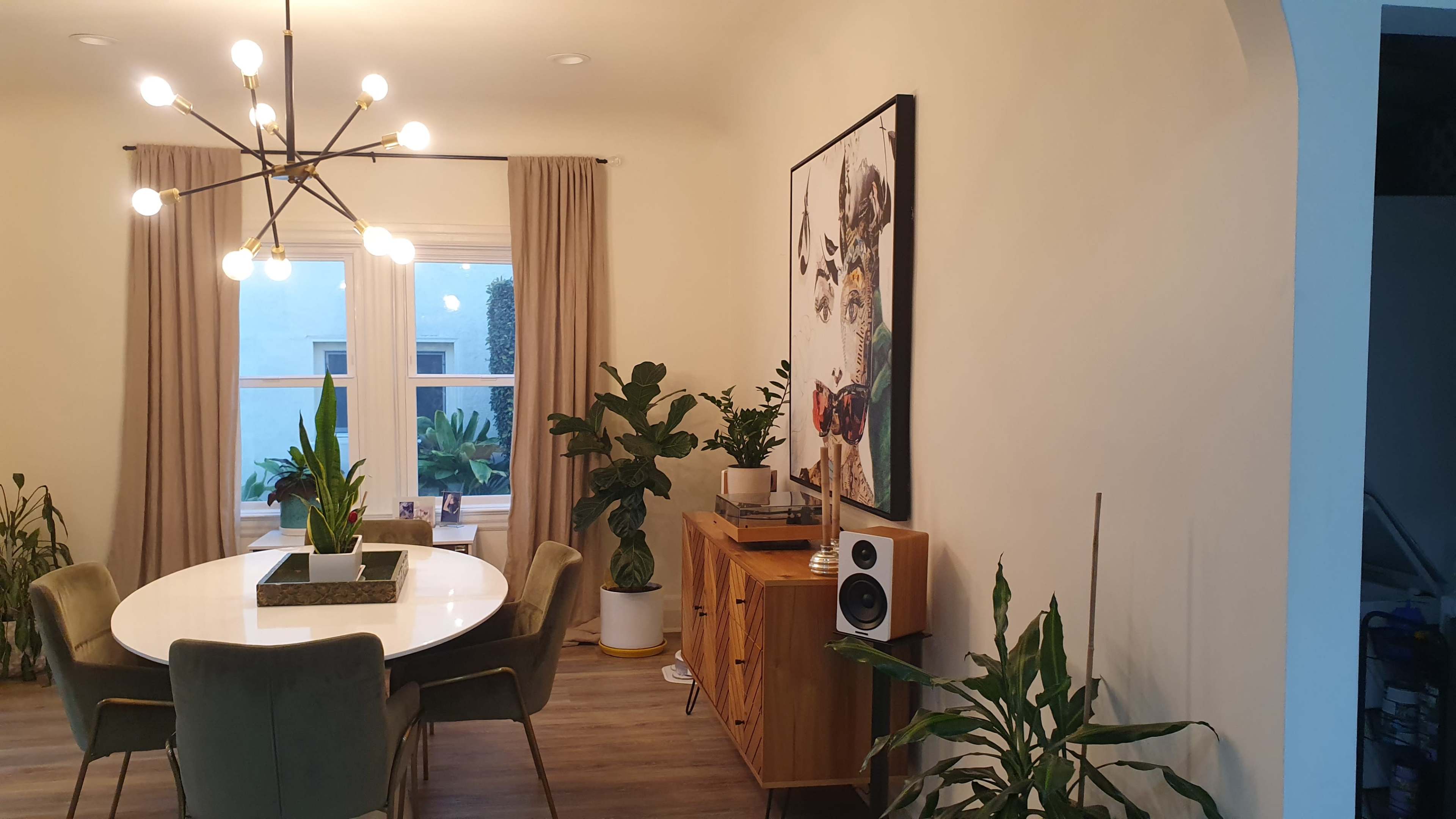 The image shows a dining area featuring a round table with green chairs, a large artwork on the wall, and various potted plants near a window.