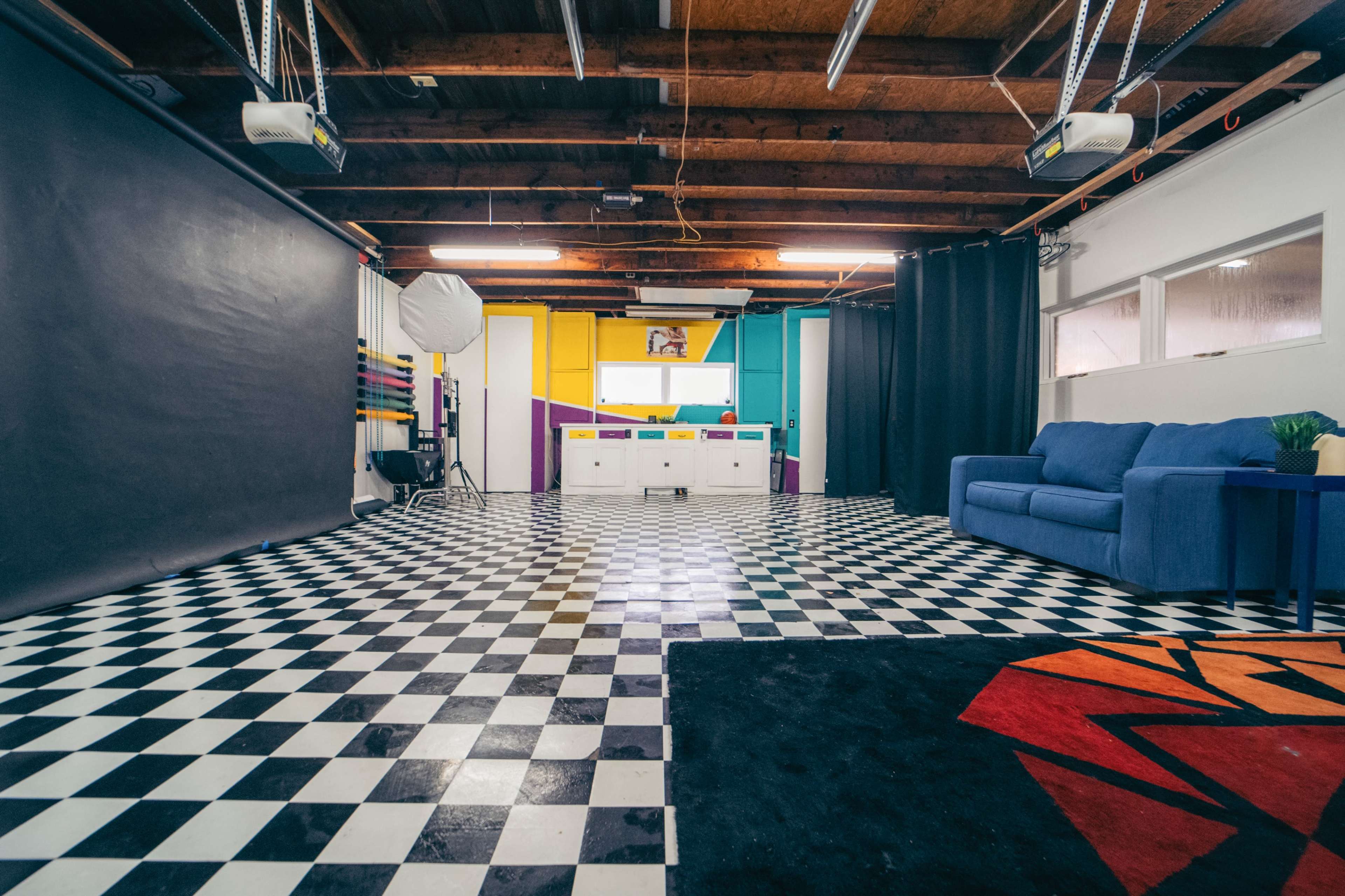The image shows a spacious studio featuring a checkered floor, a blue couch, and colorful walls with various lighting equipment.