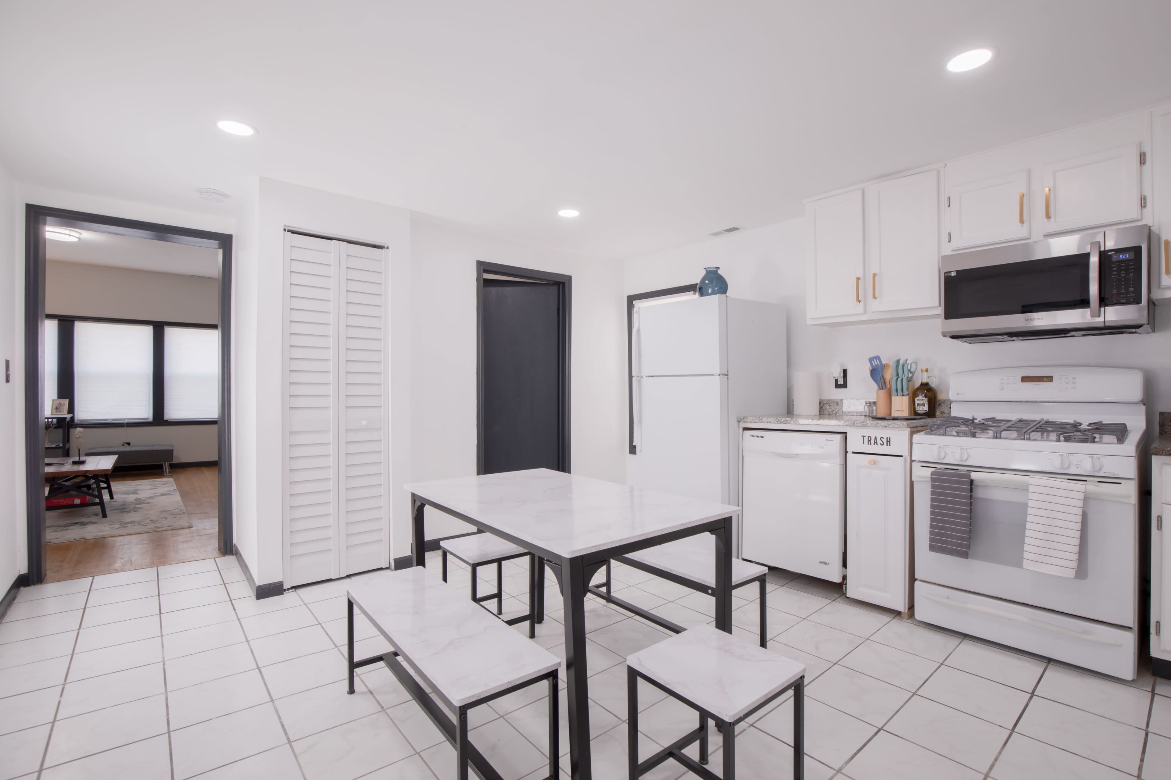 A modern kitchen features white cabinets, stainless steel appliances, and a simple dining table with benches.