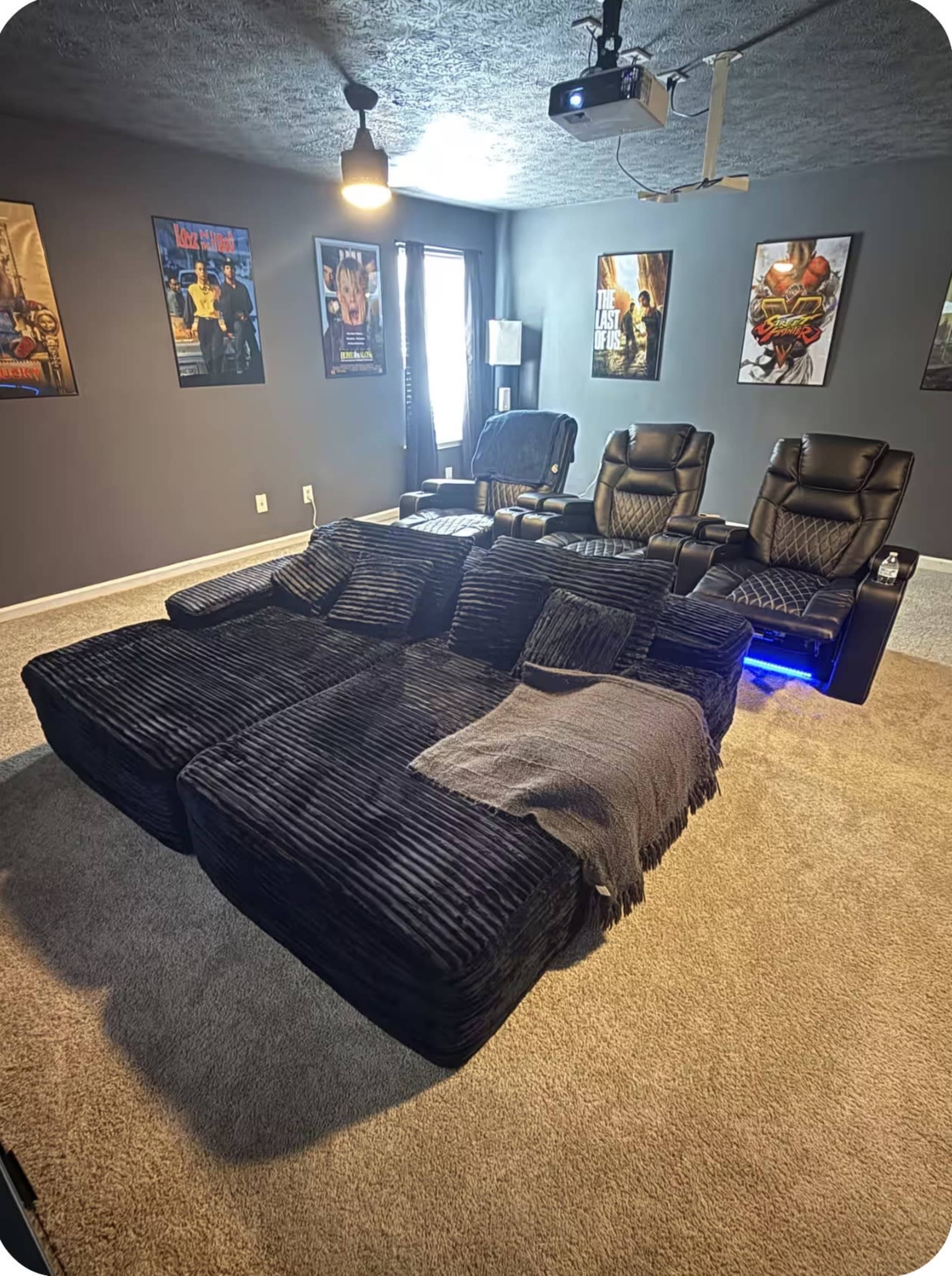 The image shows a home theater room with a large black sectional sofa, reclining chairs, a projector, and movie posters on the walls.