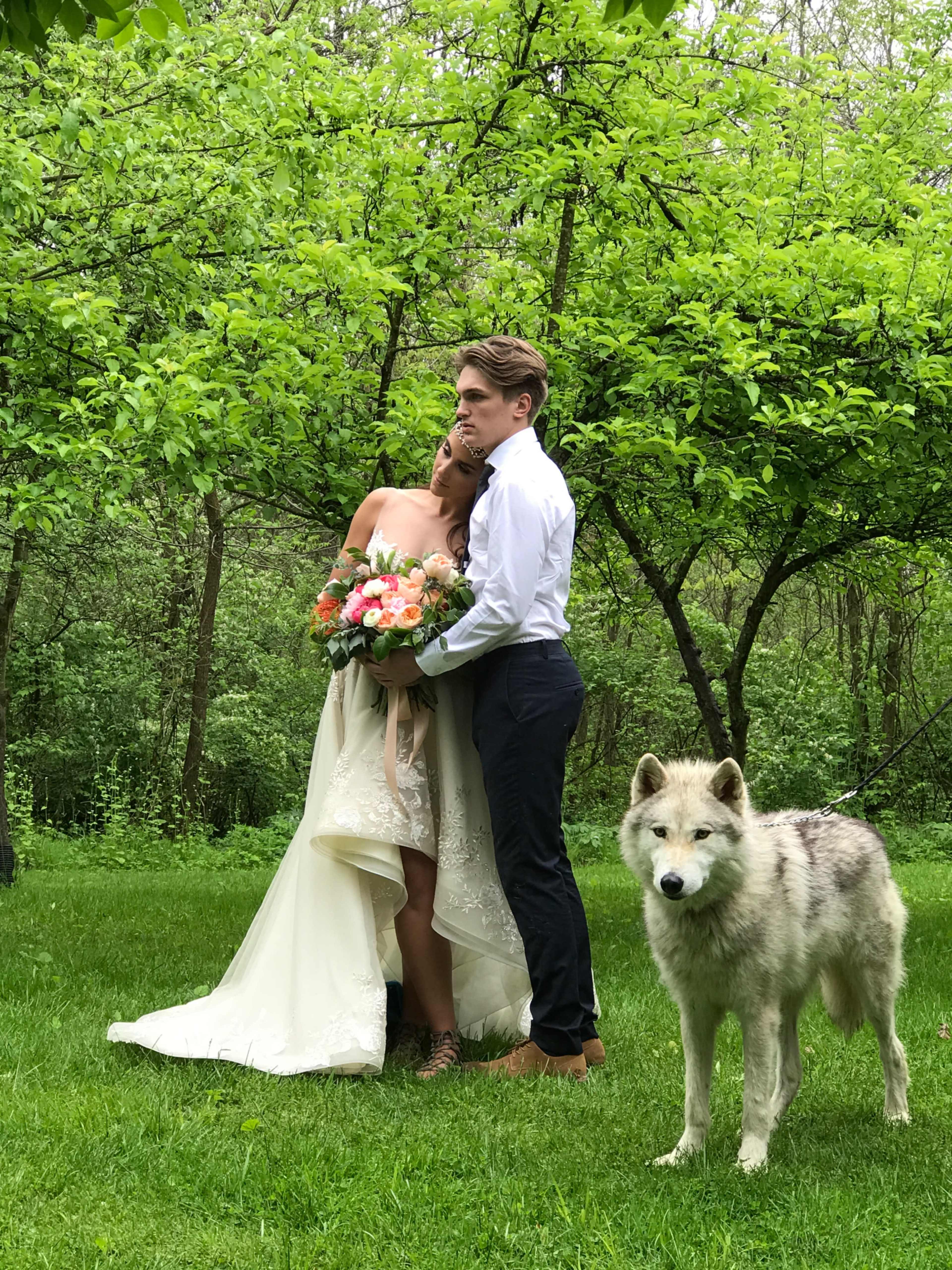 A bride holding a bouquet is embraced by a groom in a lush green setting, with a wolf standing nearby.