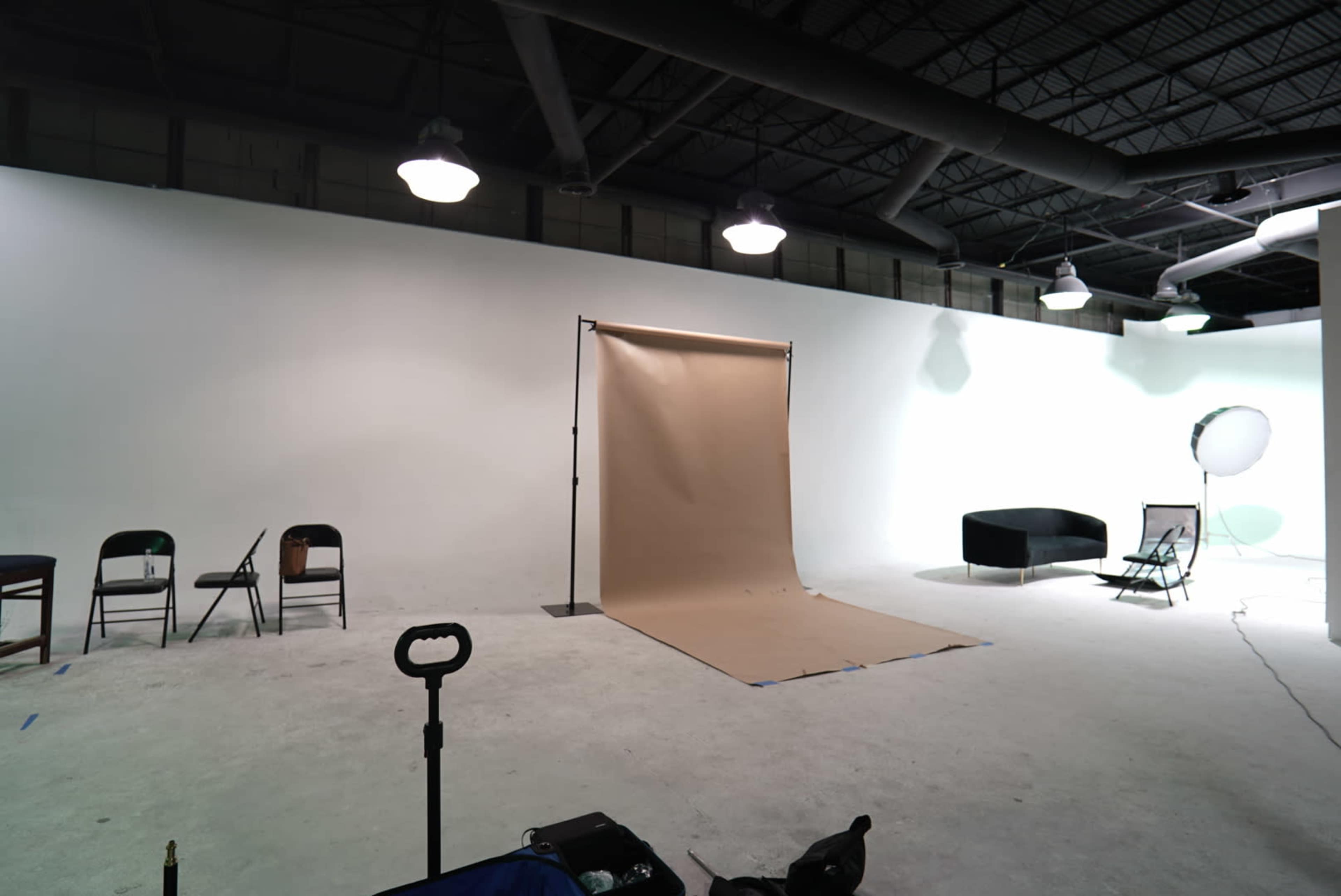 A photography studio with a beige backdrop set up on a wooden floor, surrounded by several empty chairs and a couch.