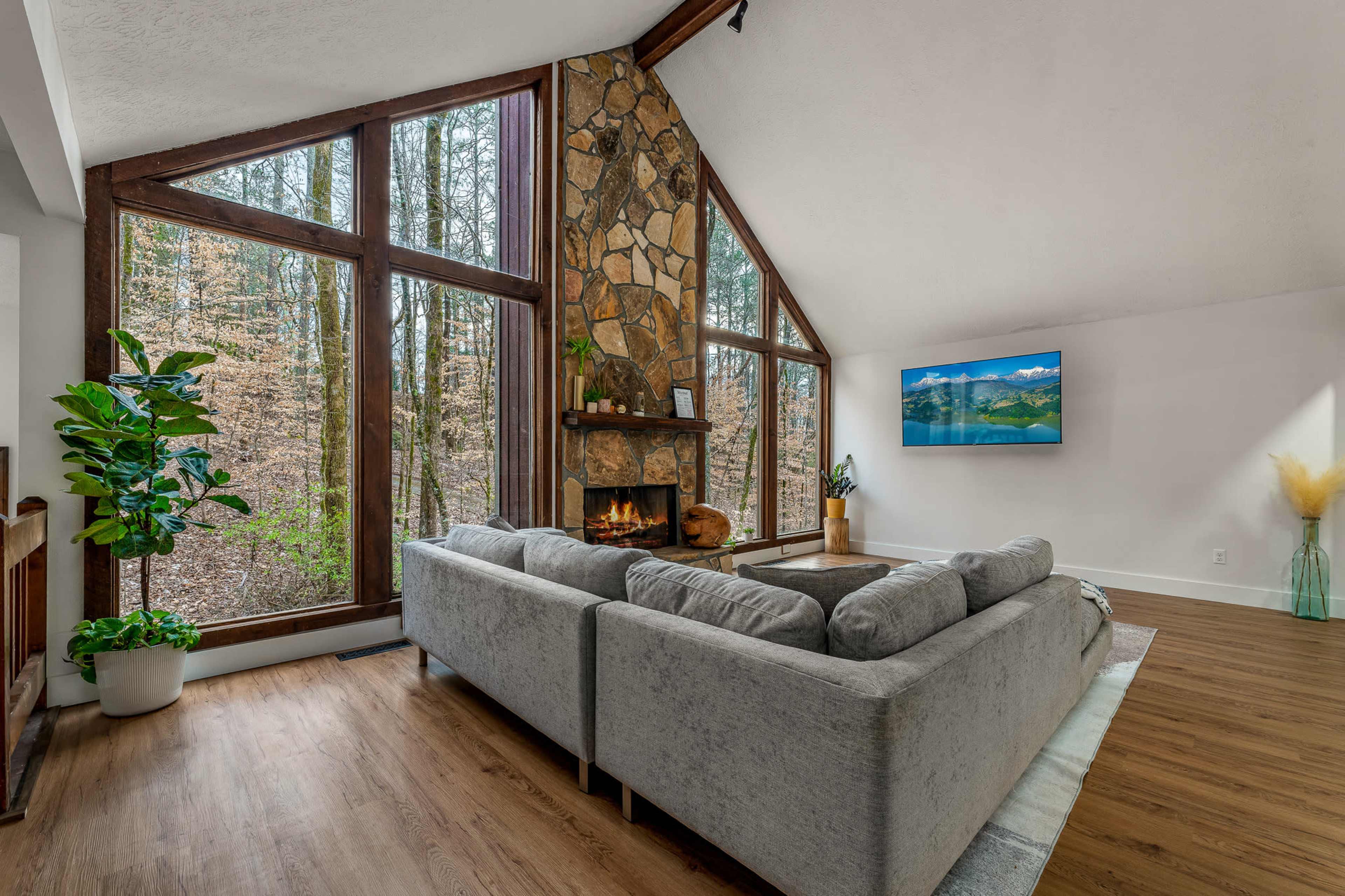 A modern living room features a stone fireplace and large windows overlooking a wooded area.