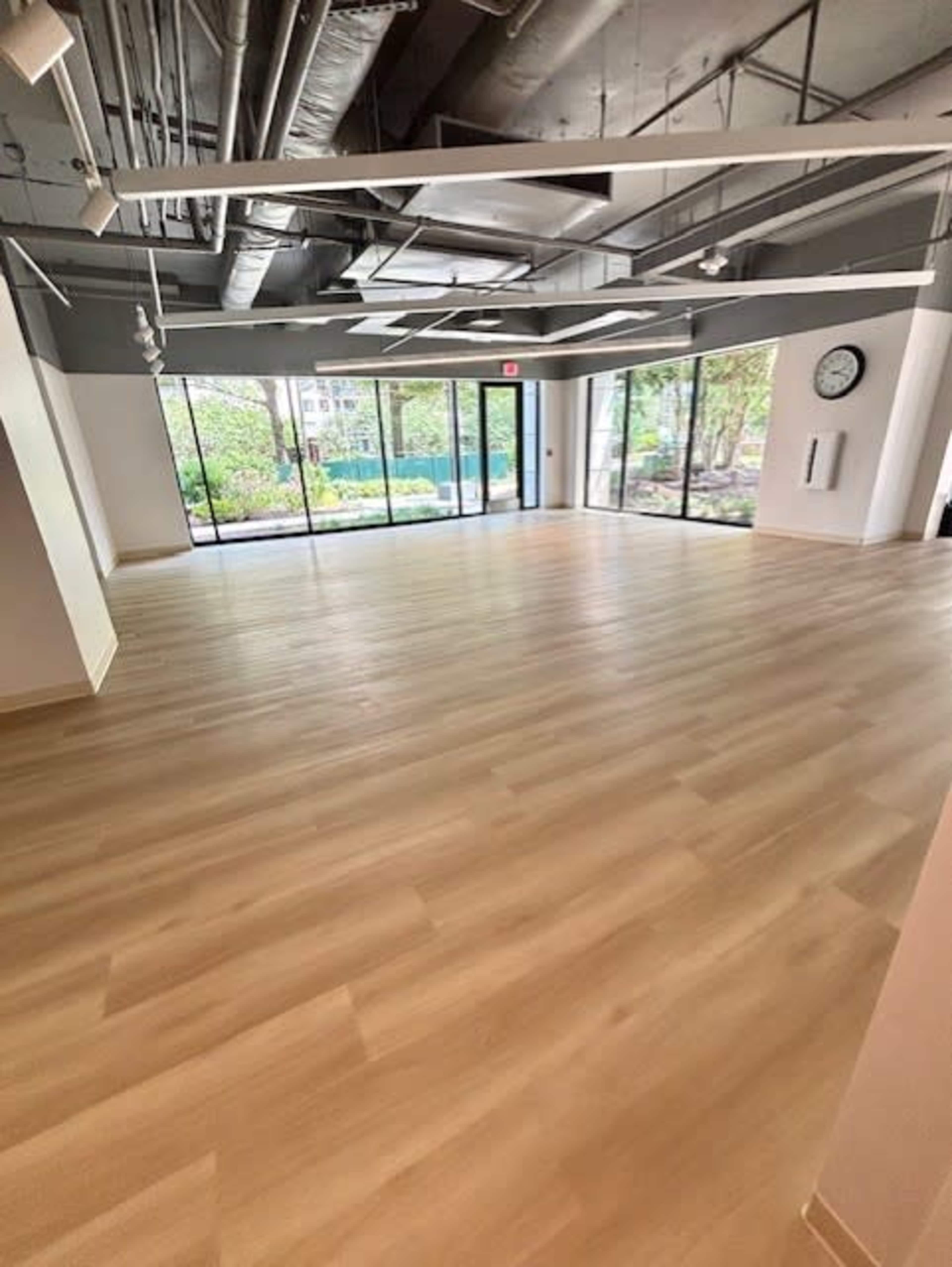 The image shows a spacious, empty room with large windows letting in natural light and displaying a view of greenery outside.