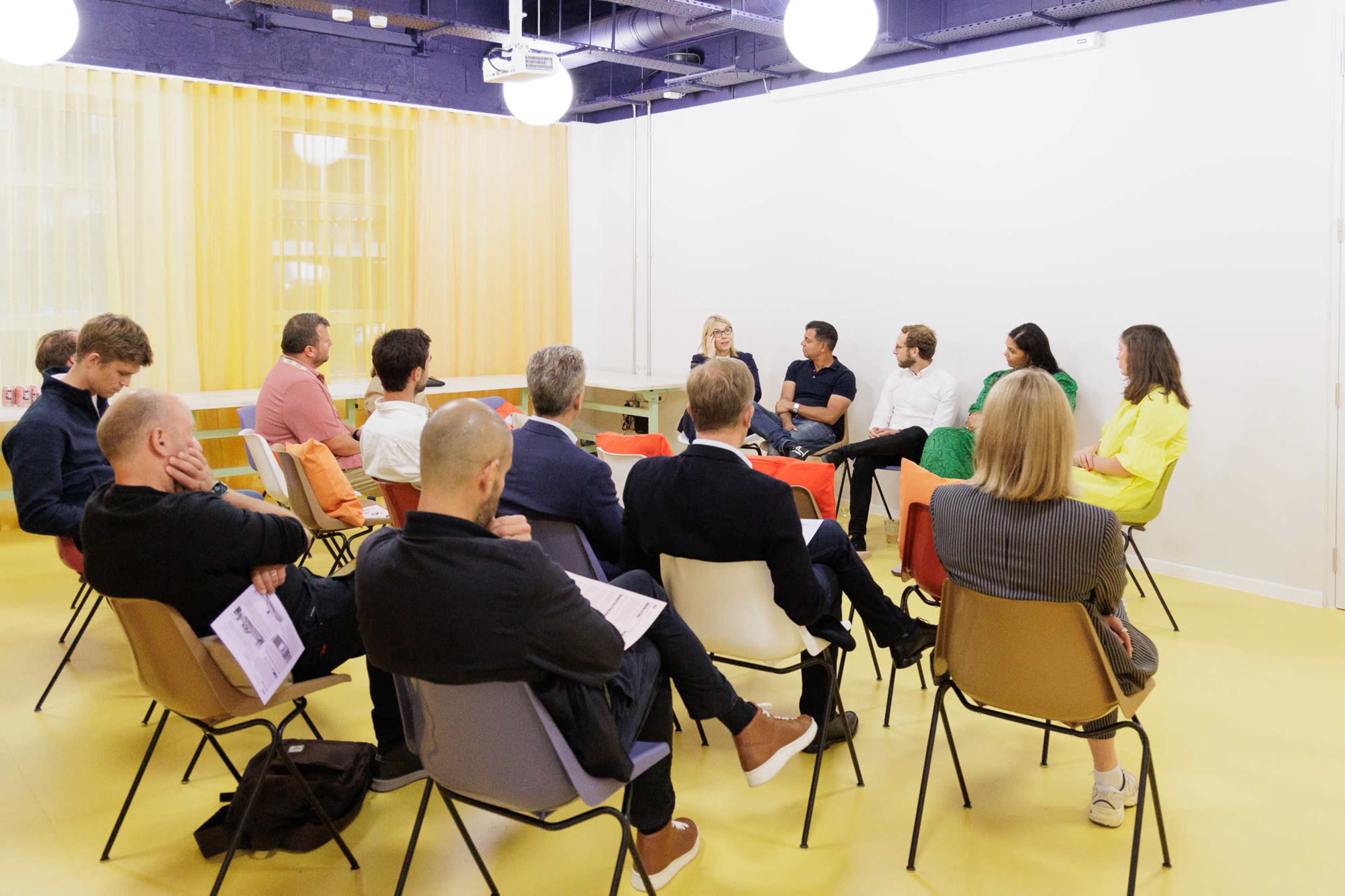 A group of people is seated in a brightly colored room, engaged in a discussion while a few individuals stand at the front.