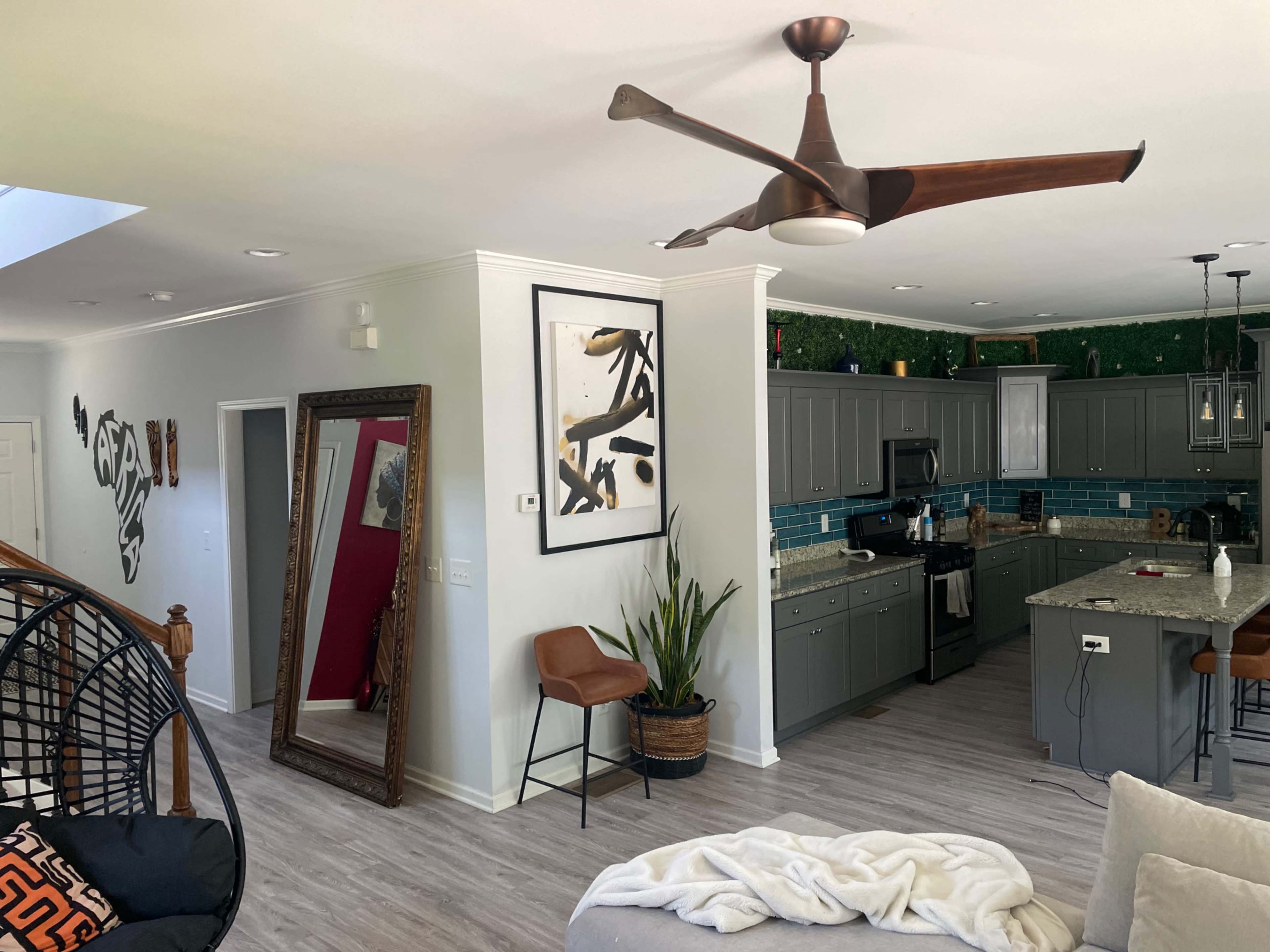 The image shows a modern open-concept living area with a kitchen, featuring gray cabinets, a large mirror, a plant, and a ceiling fan.