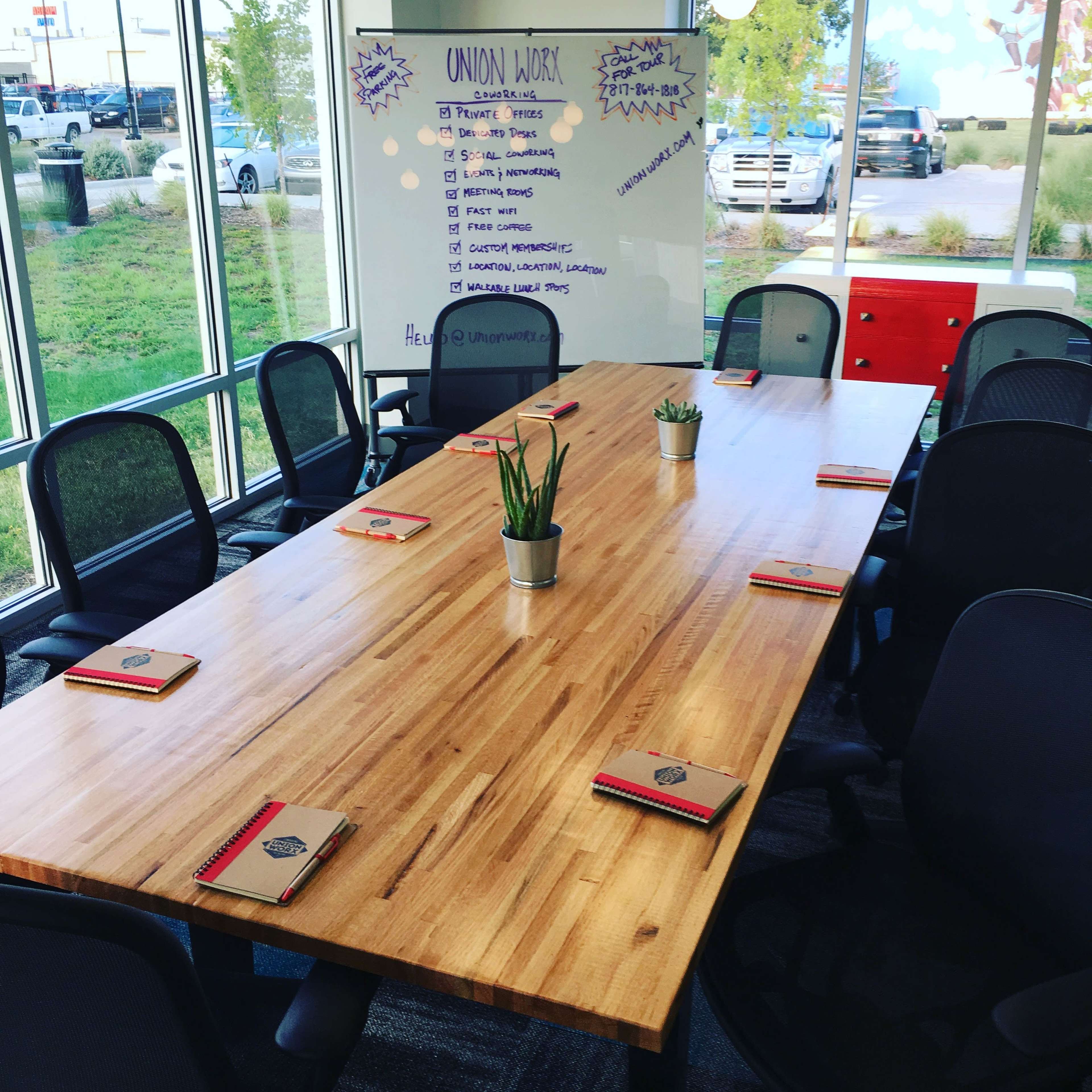 A large wooden conference table is set with notebooks and pens, surrounded by black chairs, facing a whiteboard with a list of topics written on it.